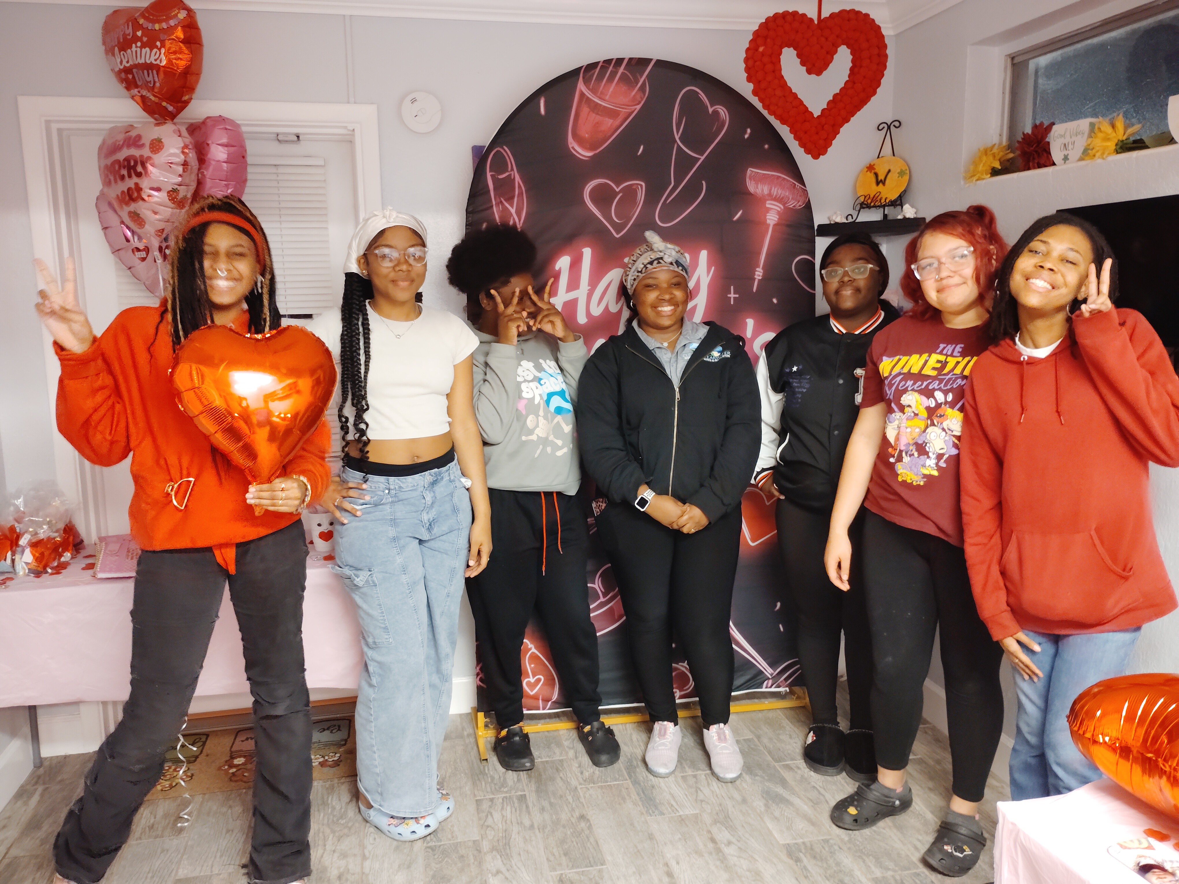 Group of seven people celebrating Valentine's Day with heart-shaped balloons and decorations, standing indoors in front of a Valentine's themed backdrop.