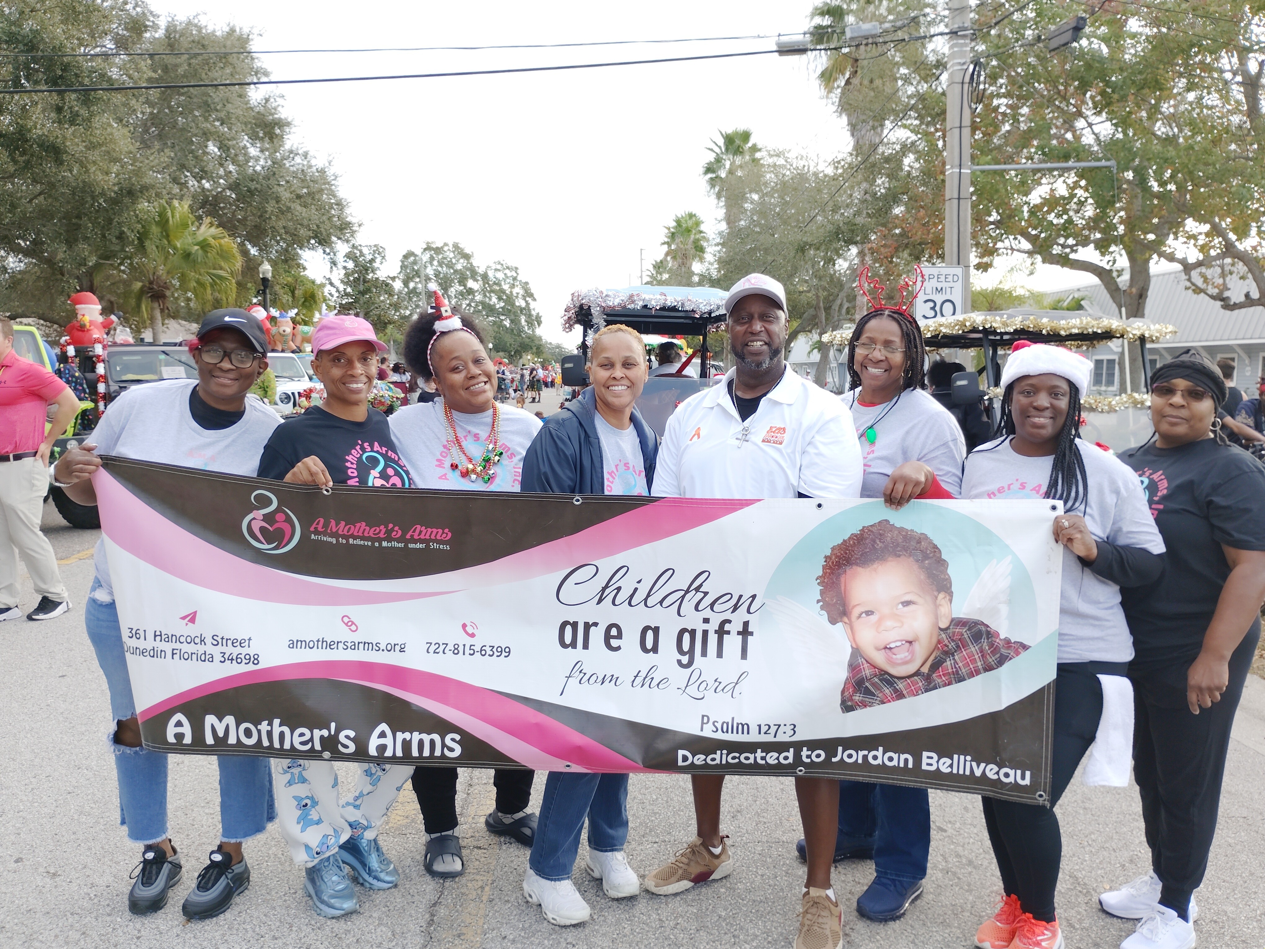 Group of people holding a banner with a child's photo, celebrating children and motherhood, outdoors with trees and cars in background.