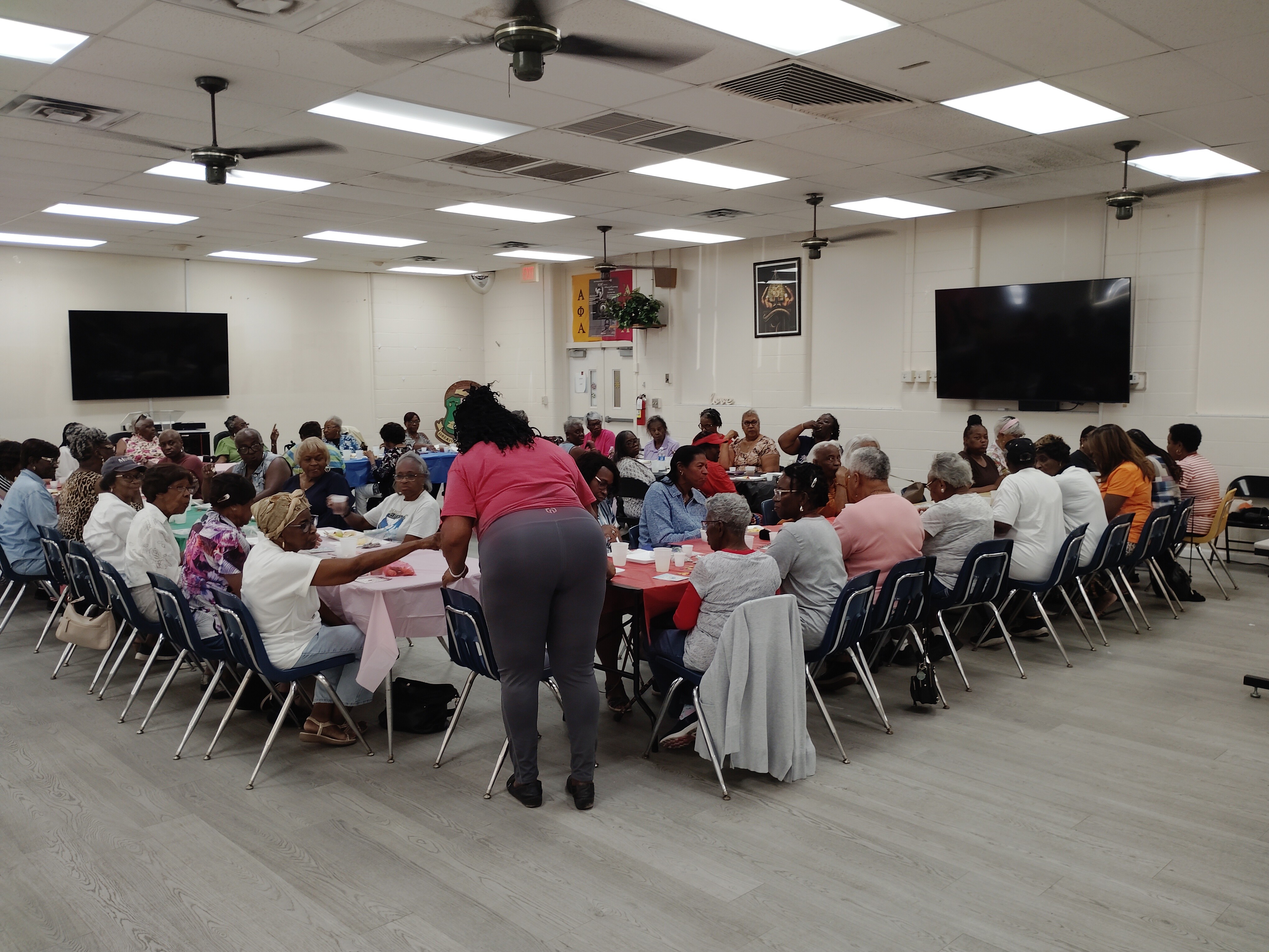 Large group of people seated around tables in a room with two large screens on the walls, ceiling fans, and fluorescent lighting.