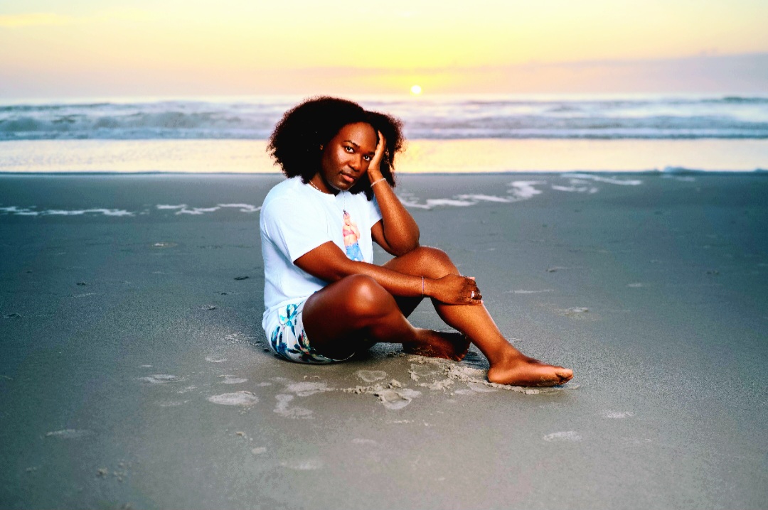 Woman sitting on the beach with ocean waves and sunset in background, resting her head on her hand, barefoot, relaxed.