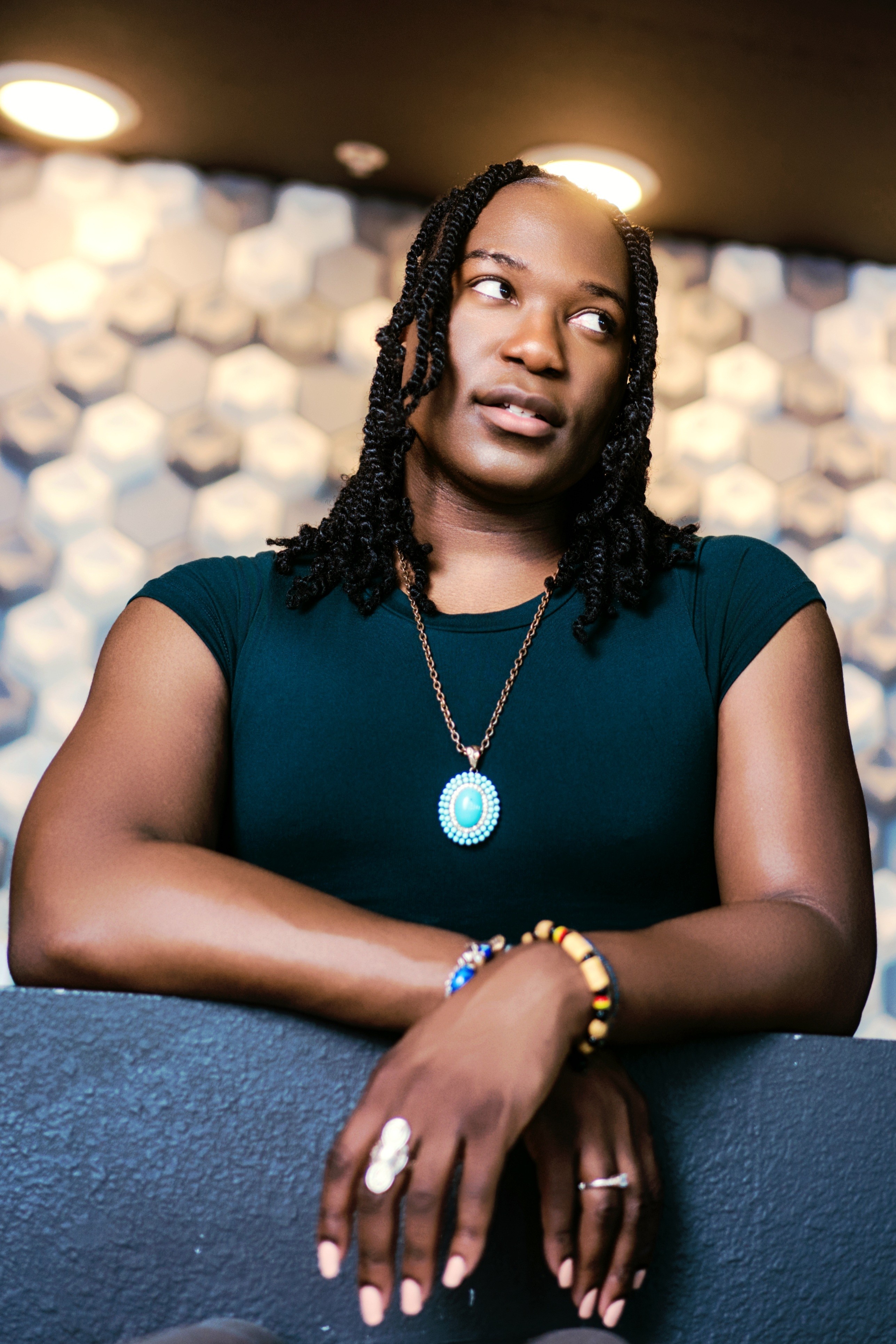 Woman with braided hair wearing a dark top and jewelry, leaning on a surface indoors, looking to the side.