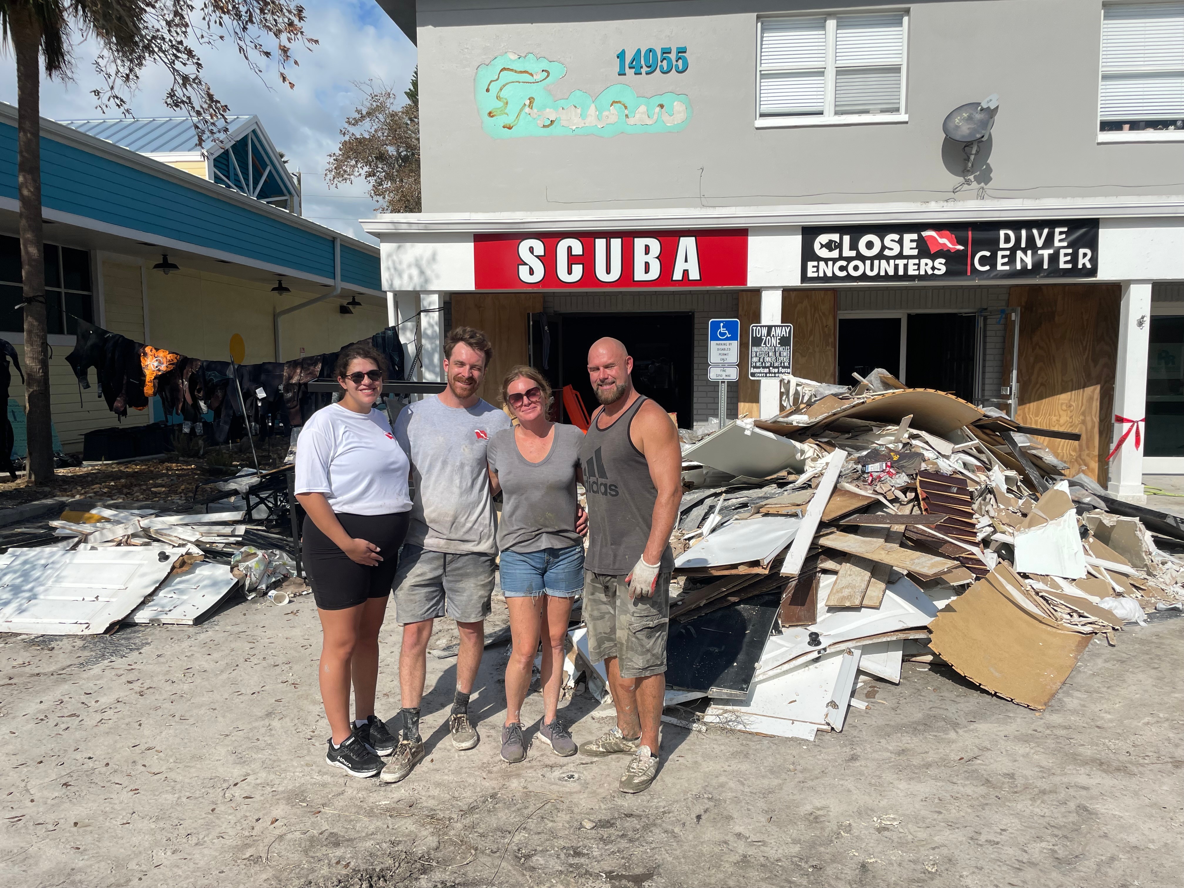 Four people stand in front of a damaged building with debris, a sign reading 'SCUBA,' and a partially destroyed structure.