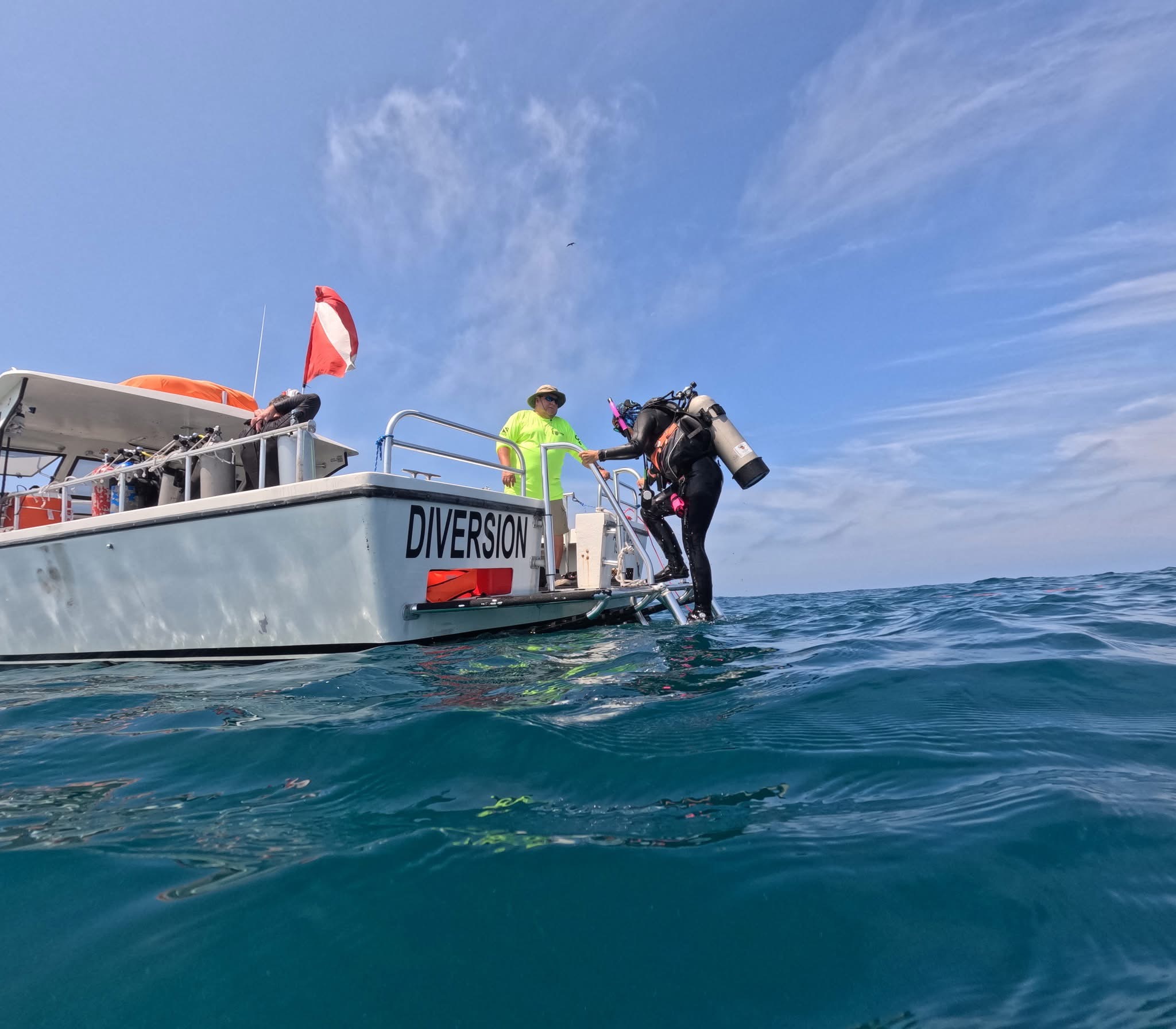 Person in wetsuit entering water from boat labeled 'DIVERSION' with crew member on deck, ocean and sky in background.