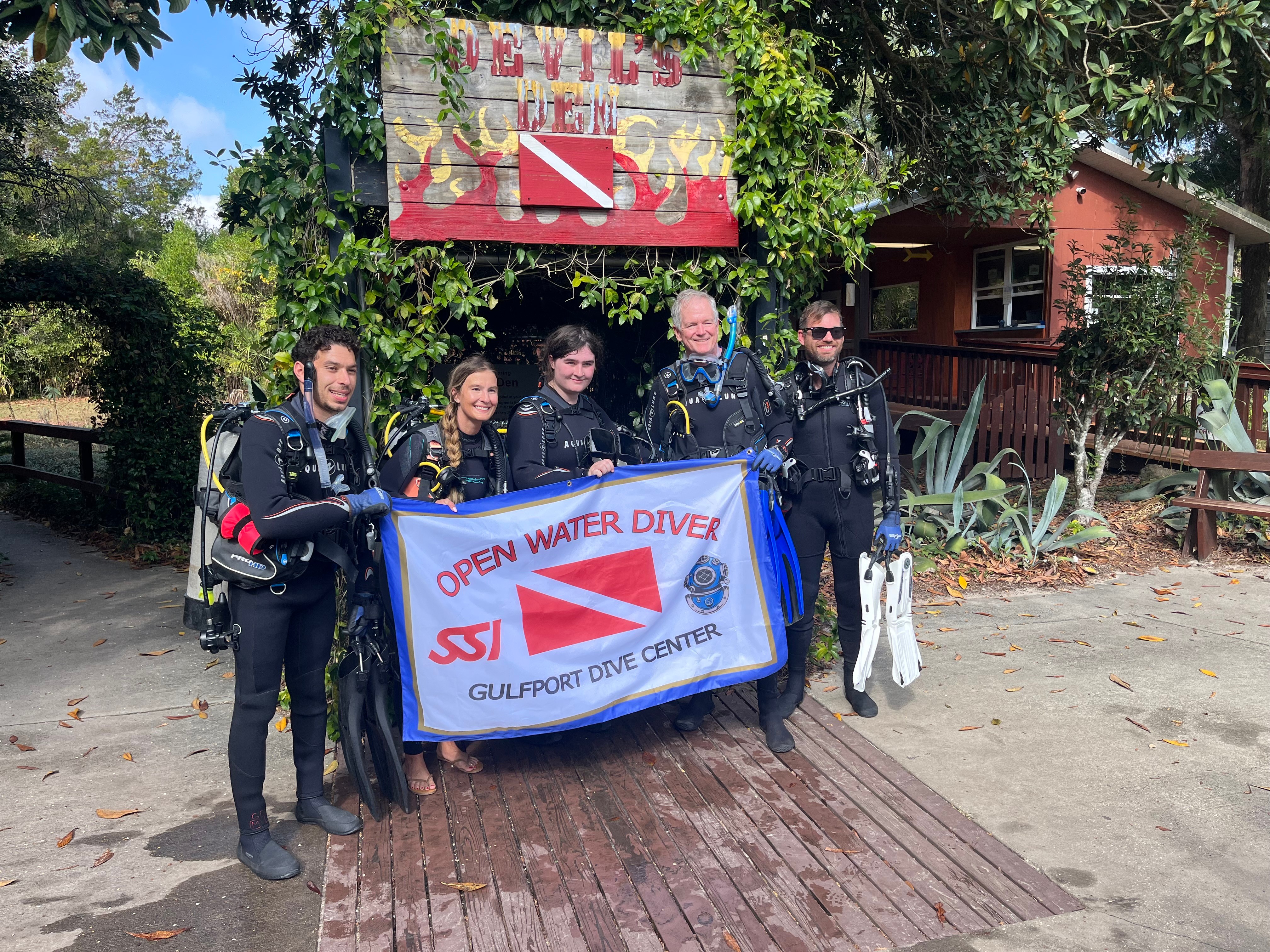 Group of five people holding a banner for open water diving, standing outdoors near trees and a building.