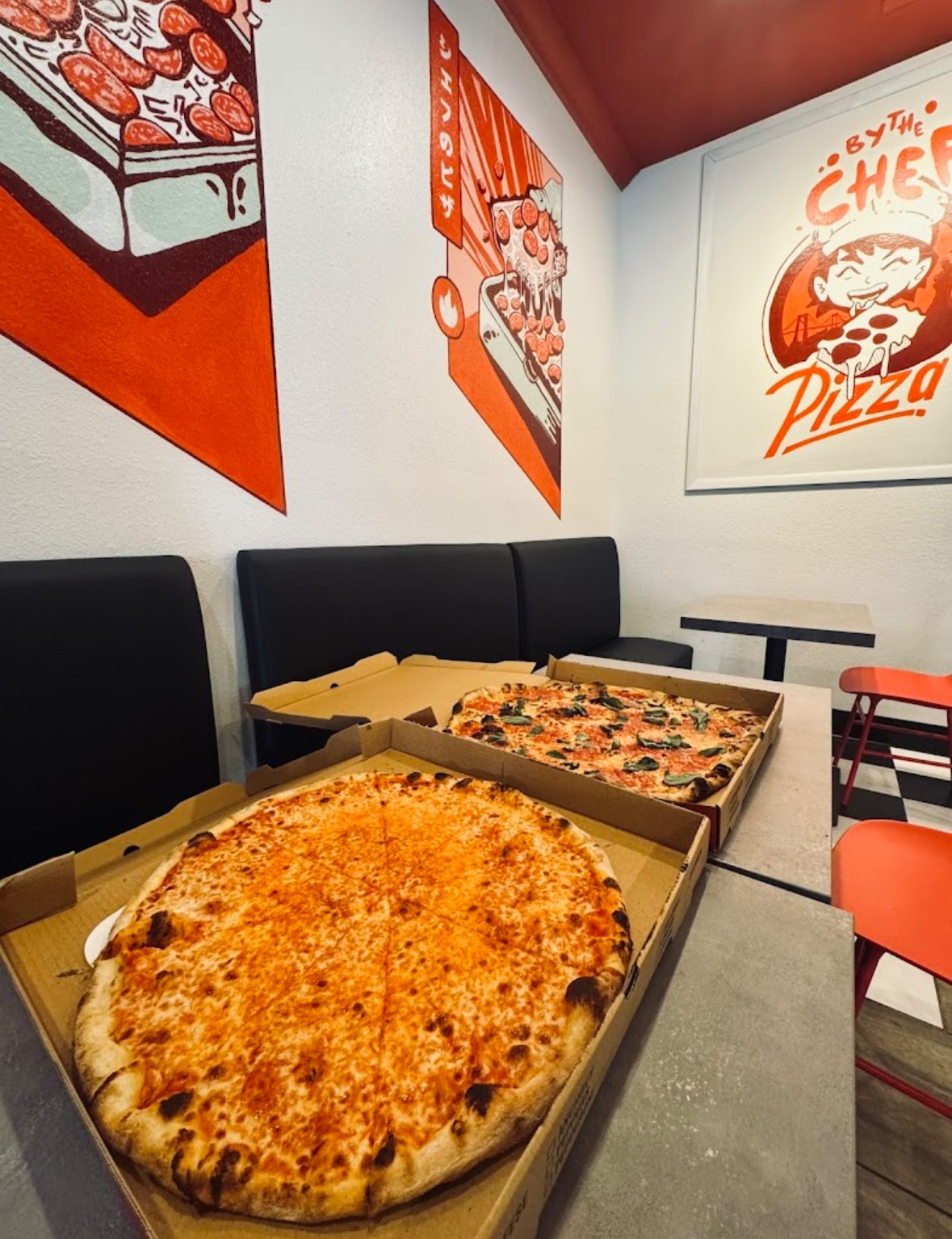 Two large pizzas in boxes on tables inside a pizzeria with colorful wall art, black and orange chairs, and a white wall.