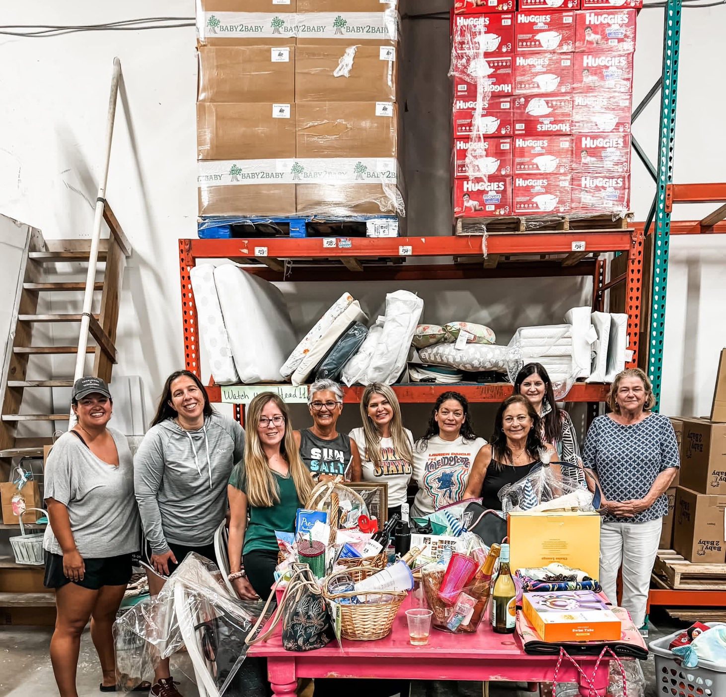 Group of nine women standing behind a table filled with bags and supplies in a warehouse.