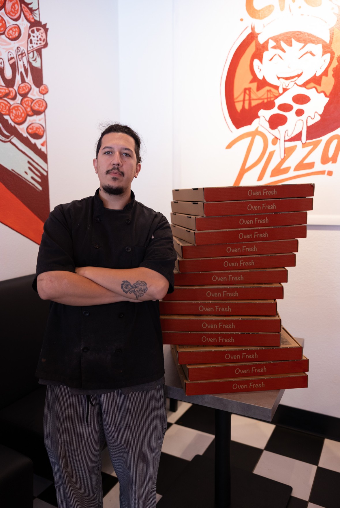 Man with a tattoo on his arm stands with arms crossed next to stacked pizza boxes inside a pizza restaurant.