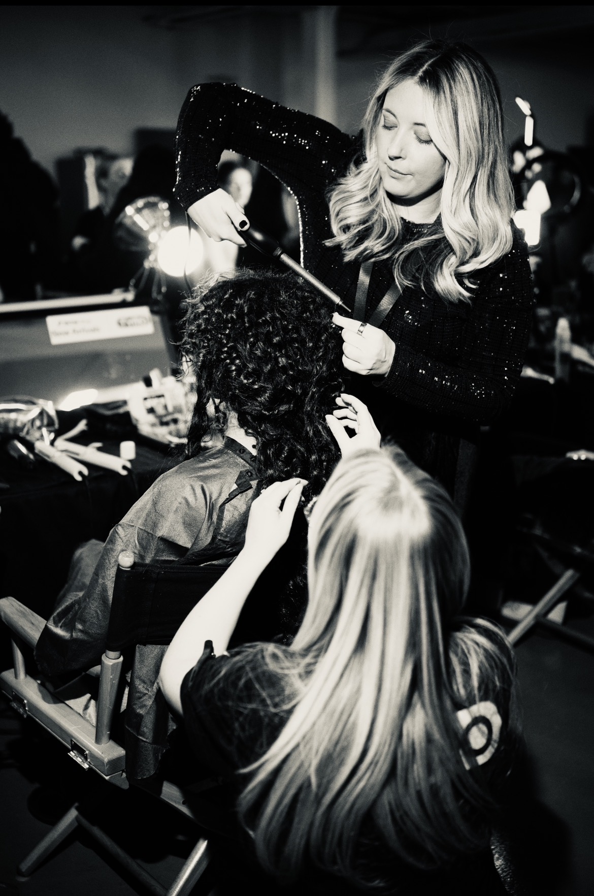 A woman with long, wavy hair applies makeup to a seated person with curly hair in a backstage setting.