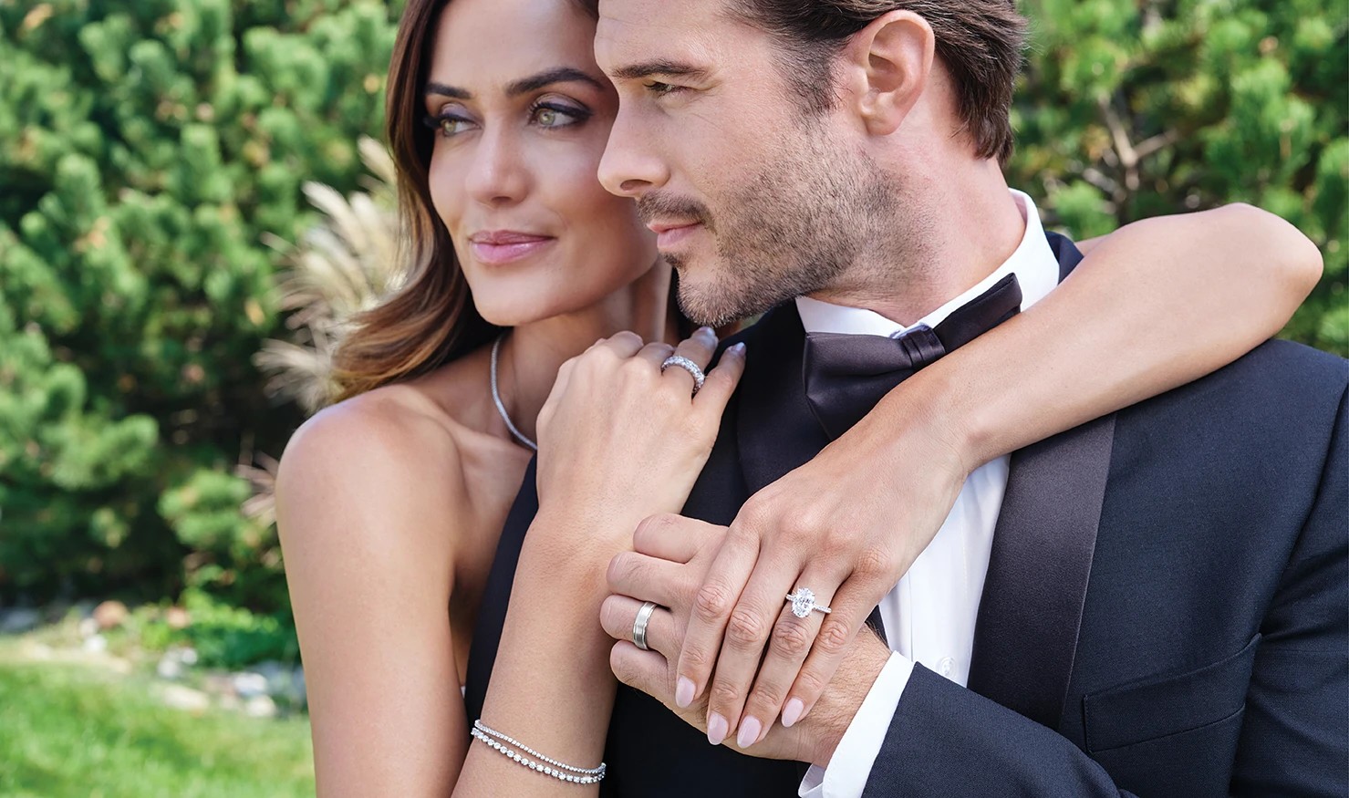 A couple dressed in formal attire, embracing outdoors with greenery in the background.