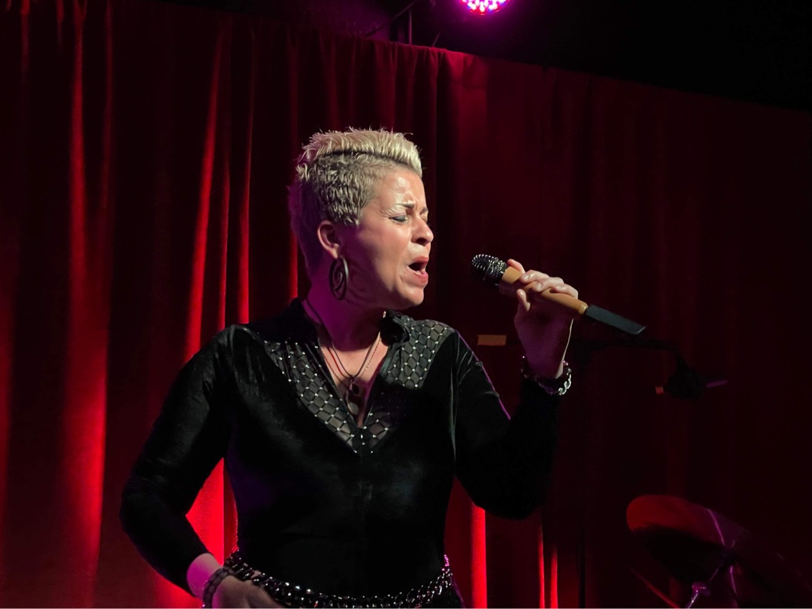 Woman singing into a microphone on stage with red curtains background.