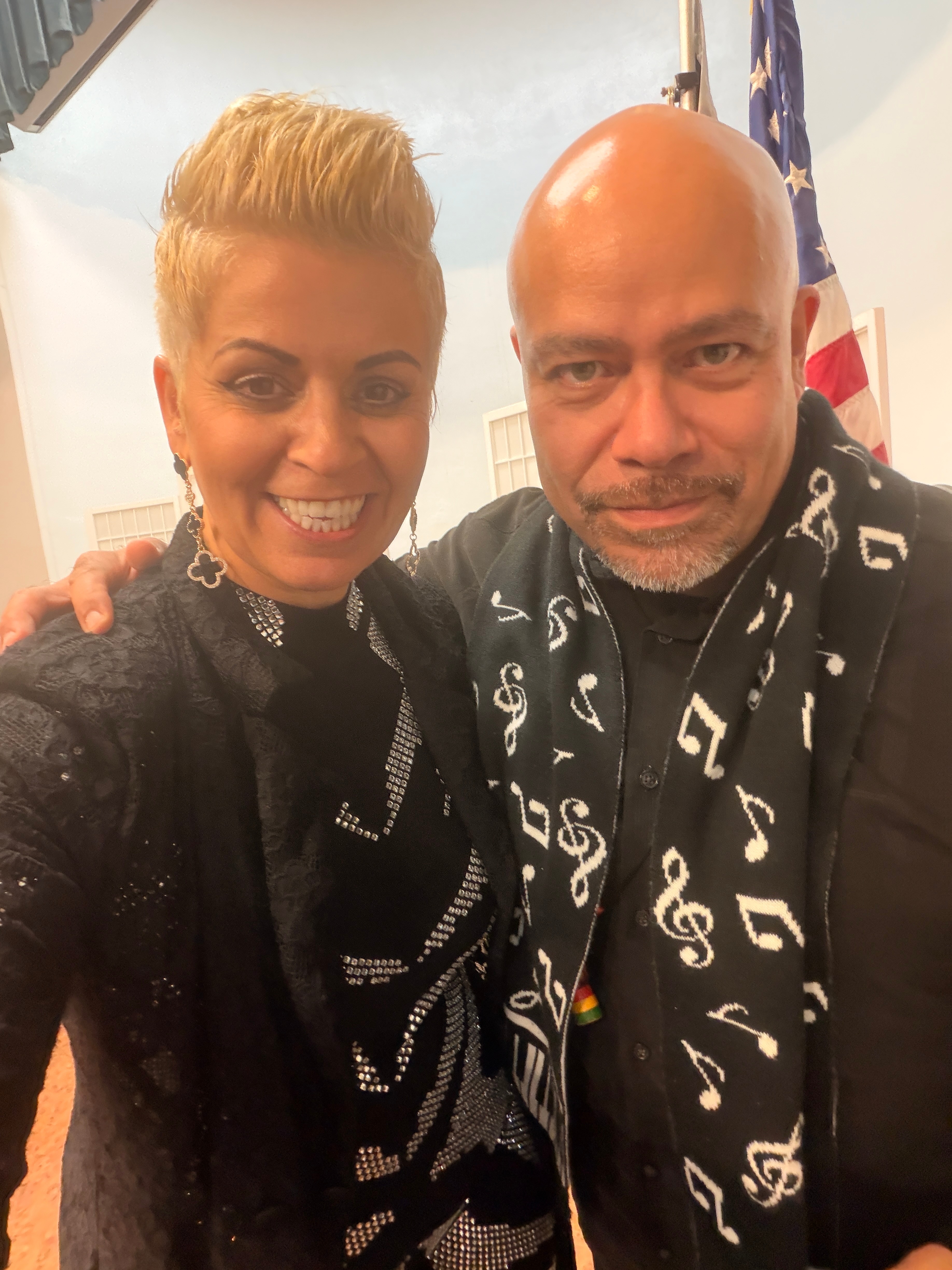 Two people smiling, one with short blonde hair and the other bald, standing close together indoors with an American flag in the background.