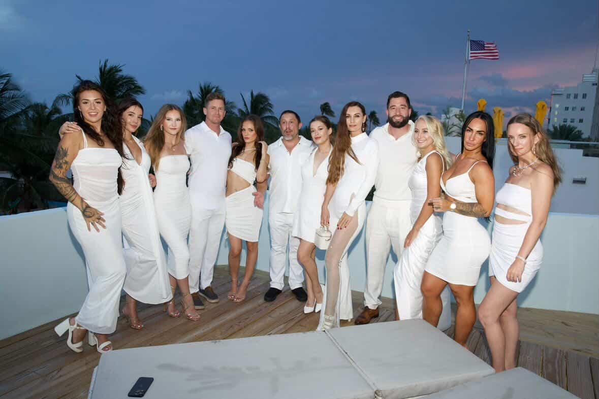 Group of people dressed in white standing on a rooftop with palm trees and an American flag in the background.