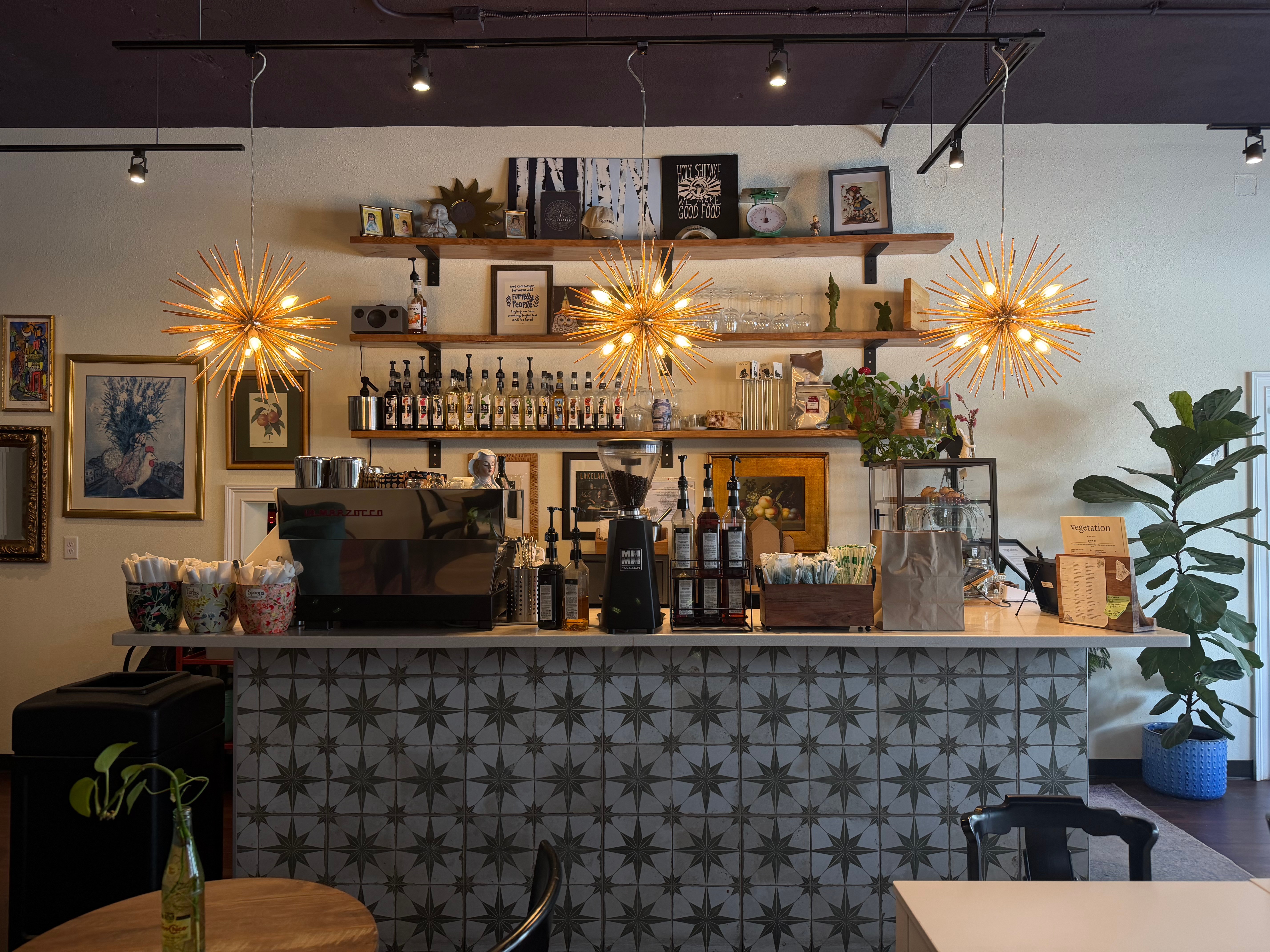 Bar counter with patterned front, shelves with bottles and decor, hanging lights, and plants in the background.