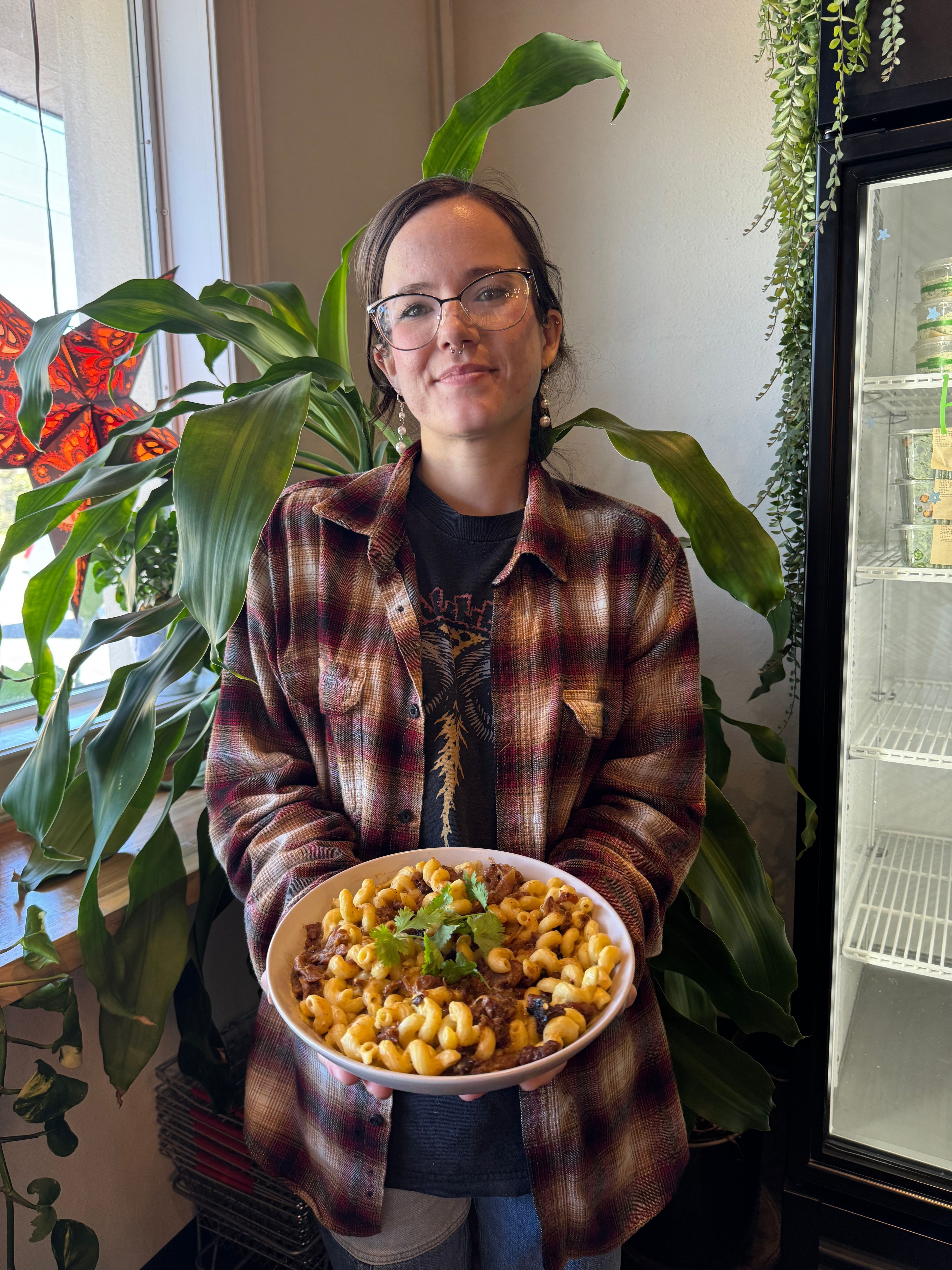 Person with glasses and long hair holding a bowl of popcorn, standing near a window with plants in the background.