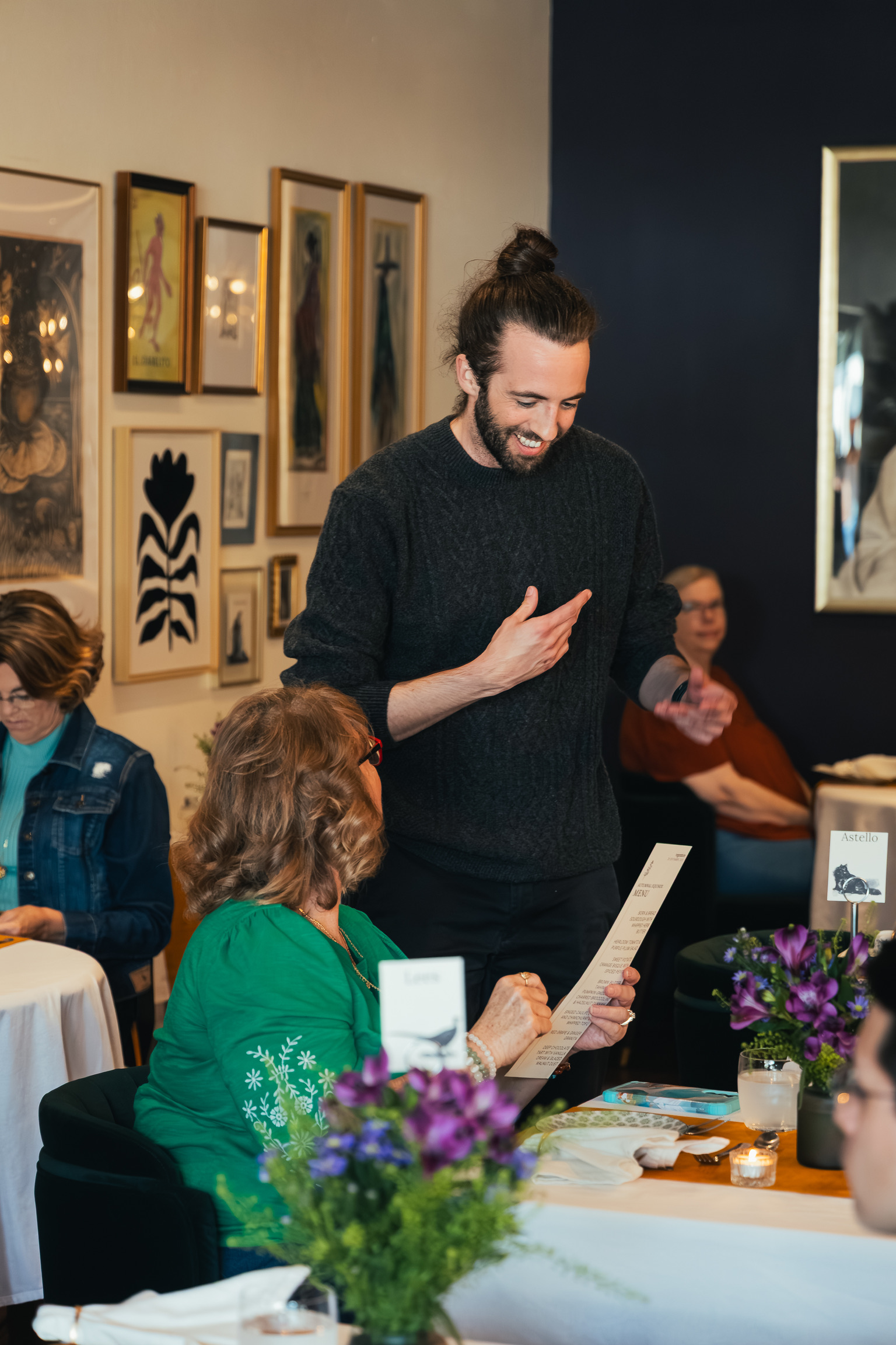 Man with a beard and long hair in a bun, smiling, standing and talking to a seated woman in a green sweater, in an art gallery setting with framed artwork on the wall. Flower arrangements on the table.