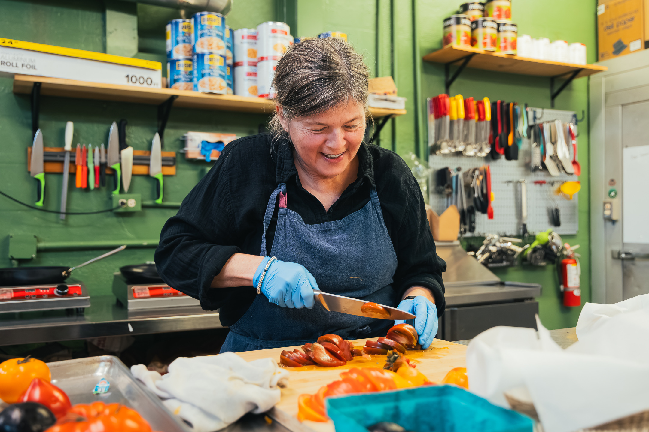 Woman slicing cooked sausages in a kitchen with green shelves and various tools, wearing a black shirt and blue gloves.