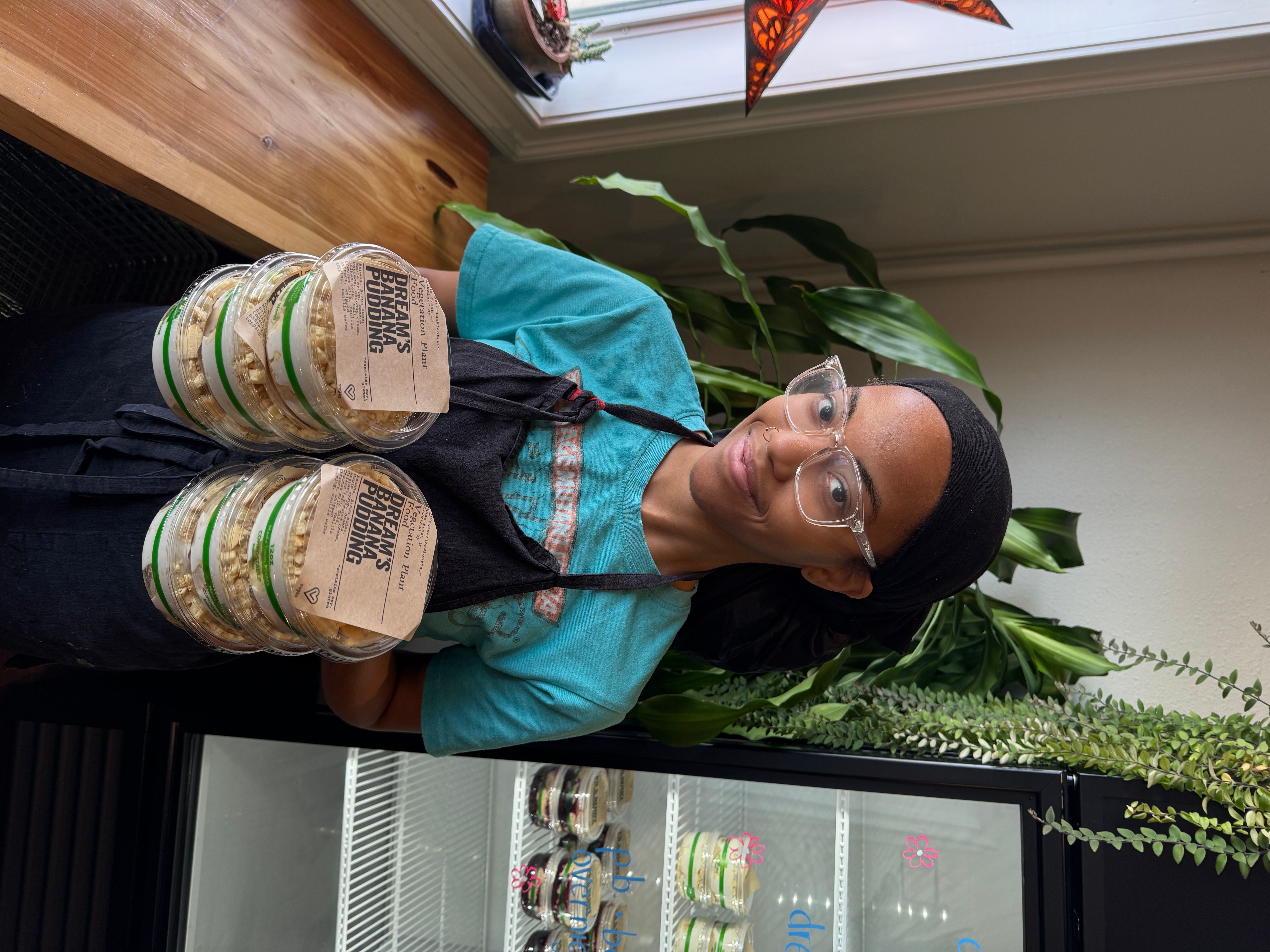 Person with glasses and black hair holding two bottles, standing indoors near a refrigerator, with plants in the background.