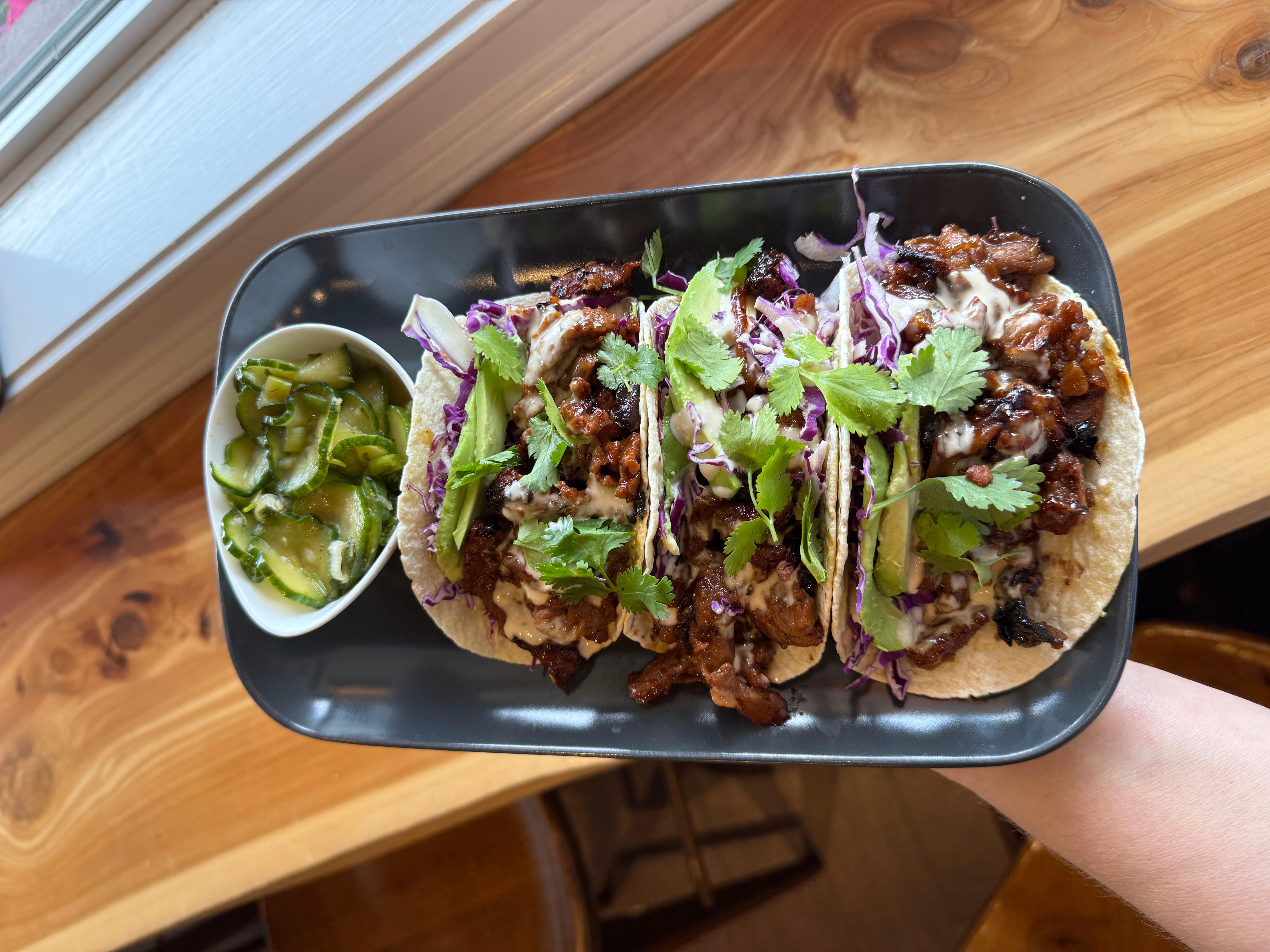 Plate with tacos topped with cilantro and purple onions, alongside a small cup of green salsa, on a wooden surface.