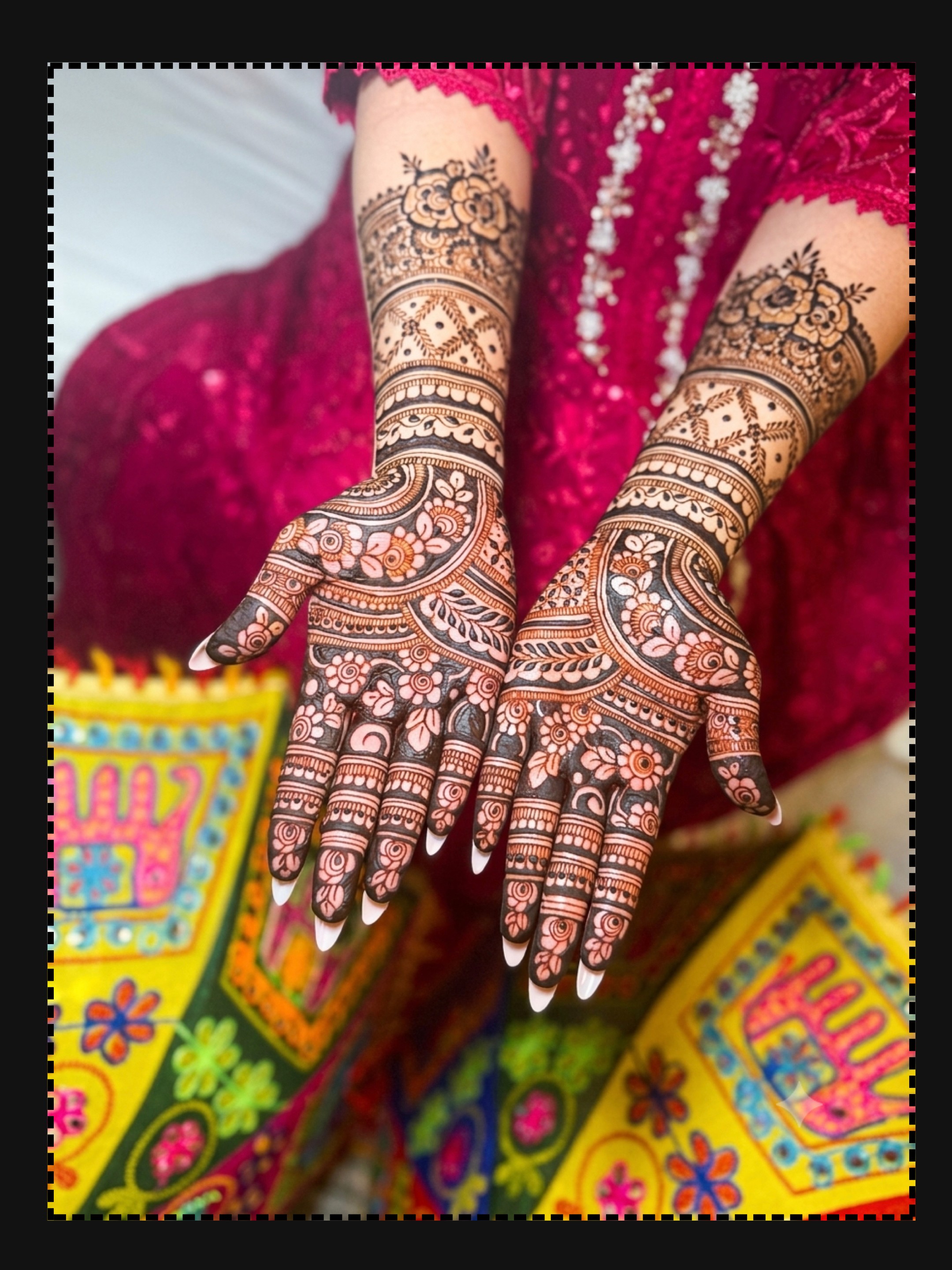 Person with decorated hands and arms, wearing colorful clothing, holding their hands together, intricate henna designs visible.