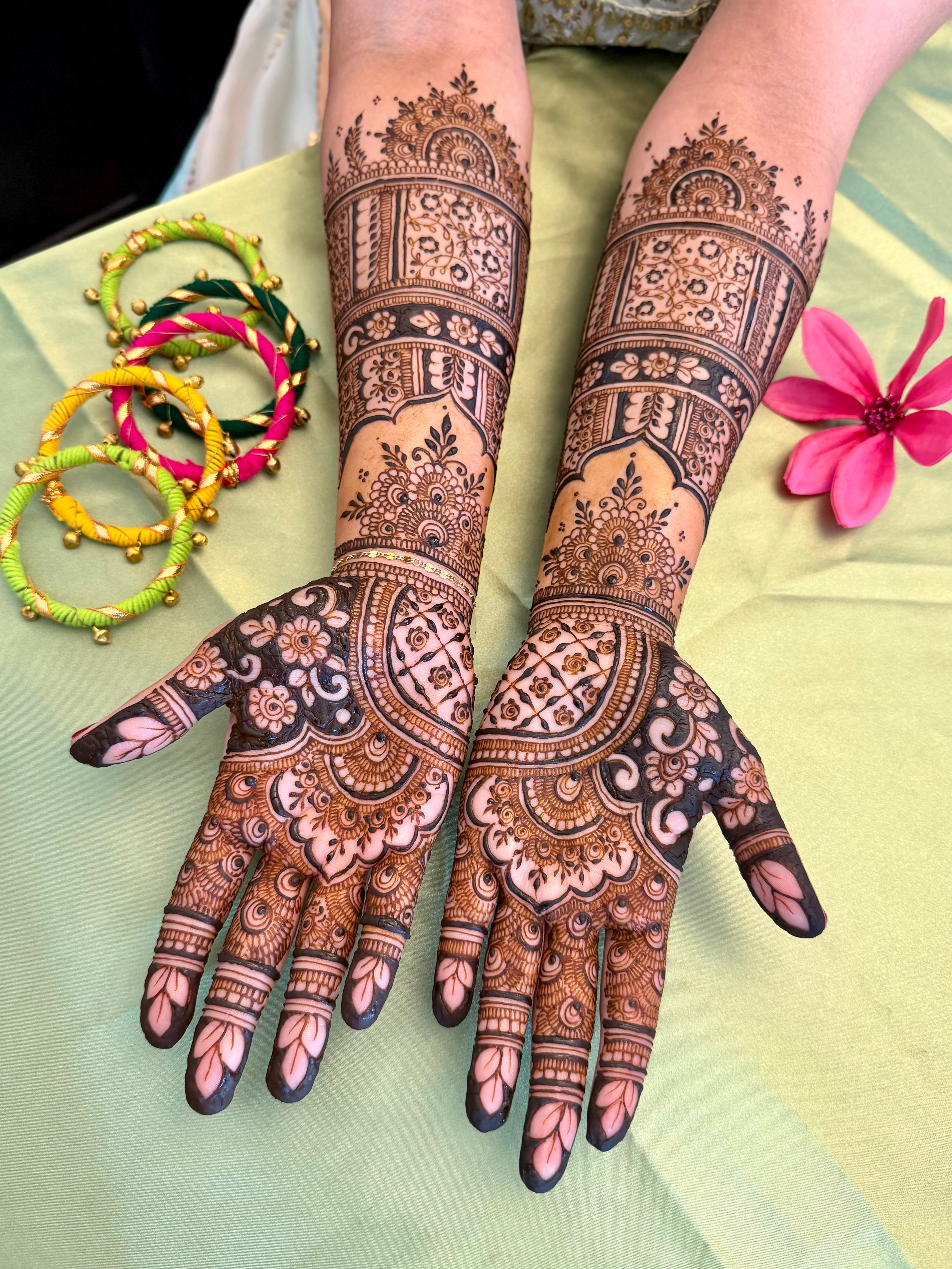 Two hands with intricate henna designs, colorful bangles, and a pink flower on a green surface.