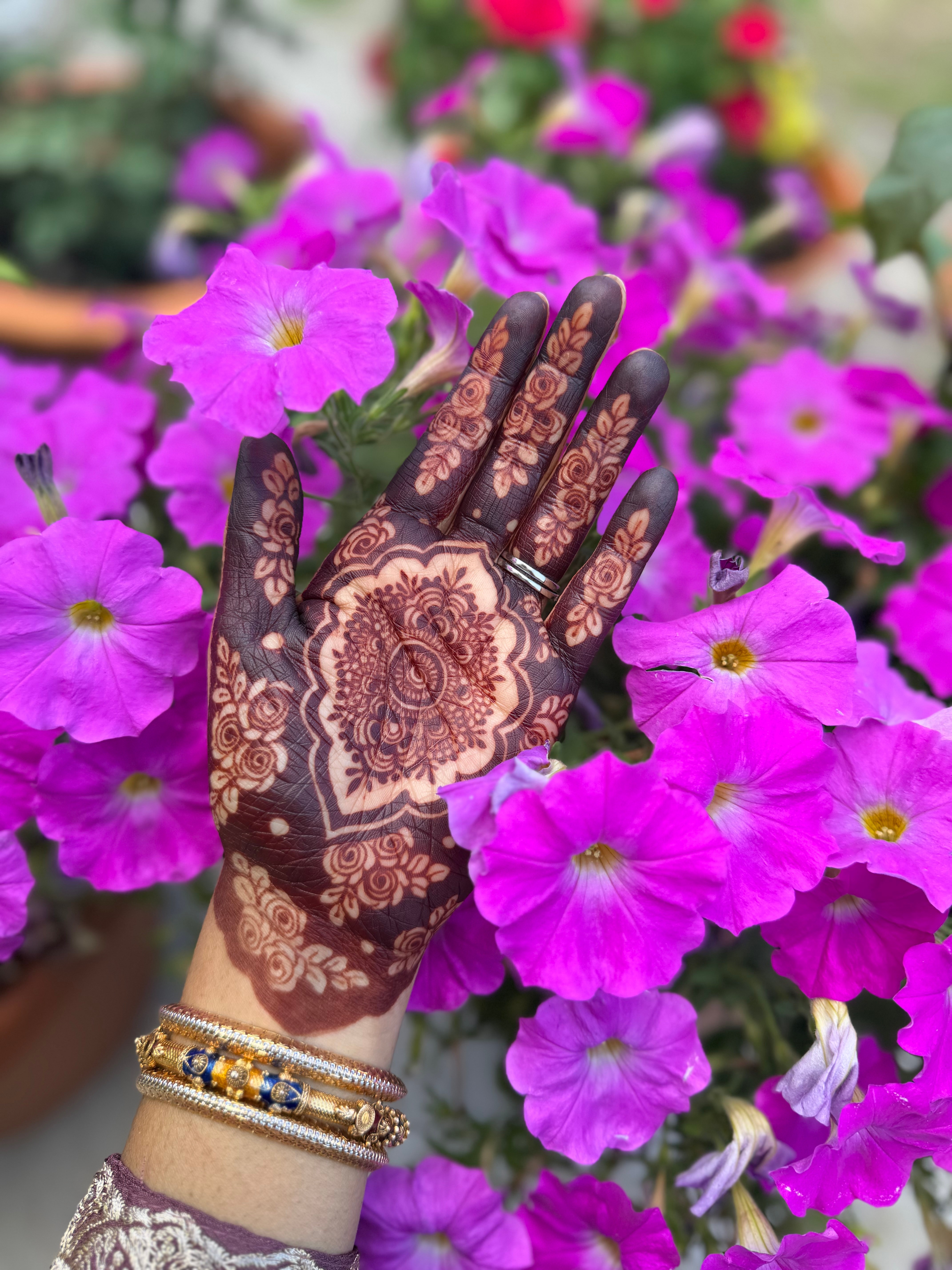 Hand with henna designs surrounded by pink and purple flowers, wearing bracelets, in a garden setting.