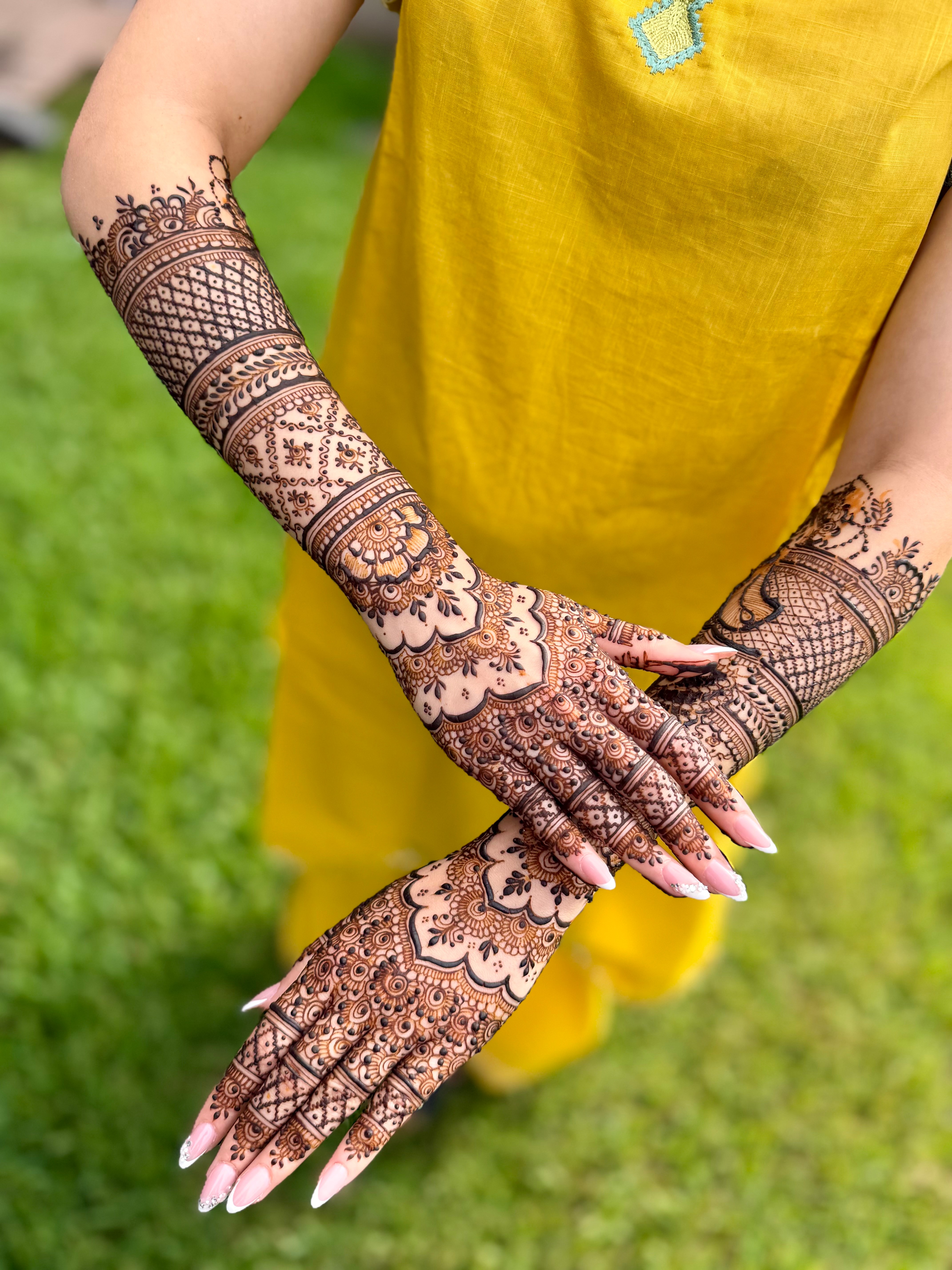 Person with intricate henna designs on arms and hands, wearing a yellow dress, standing outdoors on green grass.