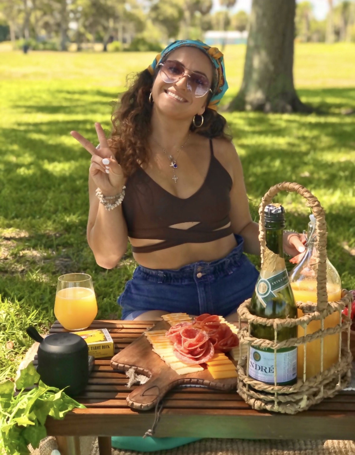 Woman with sunglasses and headphones smiling outdoors, making a peace sign, with a picnic setup including drinks and food on a table.