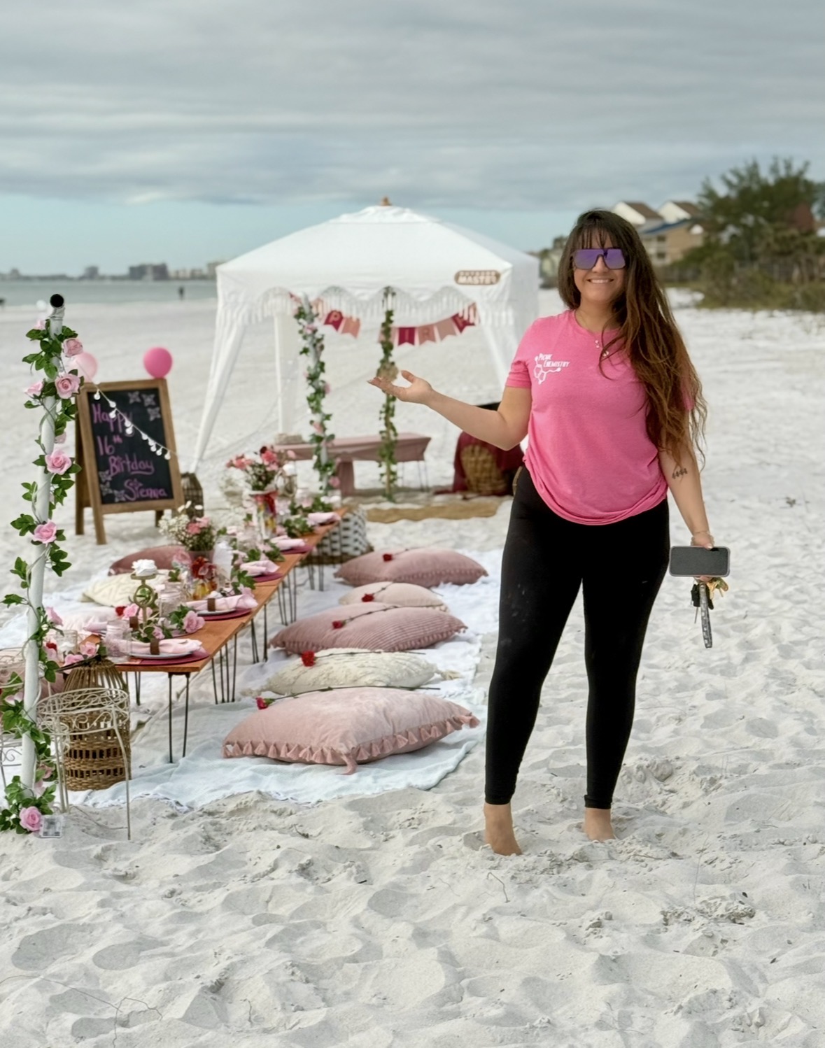Woman in pink shirt and black pants standing on beach near decorated table with flowers, pillows, and umbrella, holding a phone.
