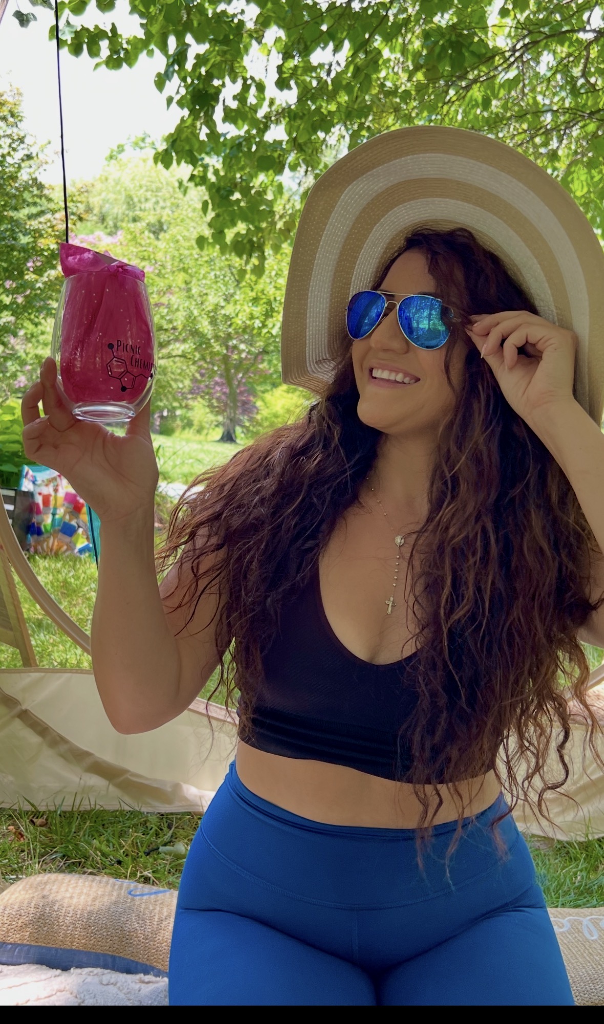 Woman outdoors wearing a large hat and sunglasses, holding a pink drink, smiling, with trees in the background.
