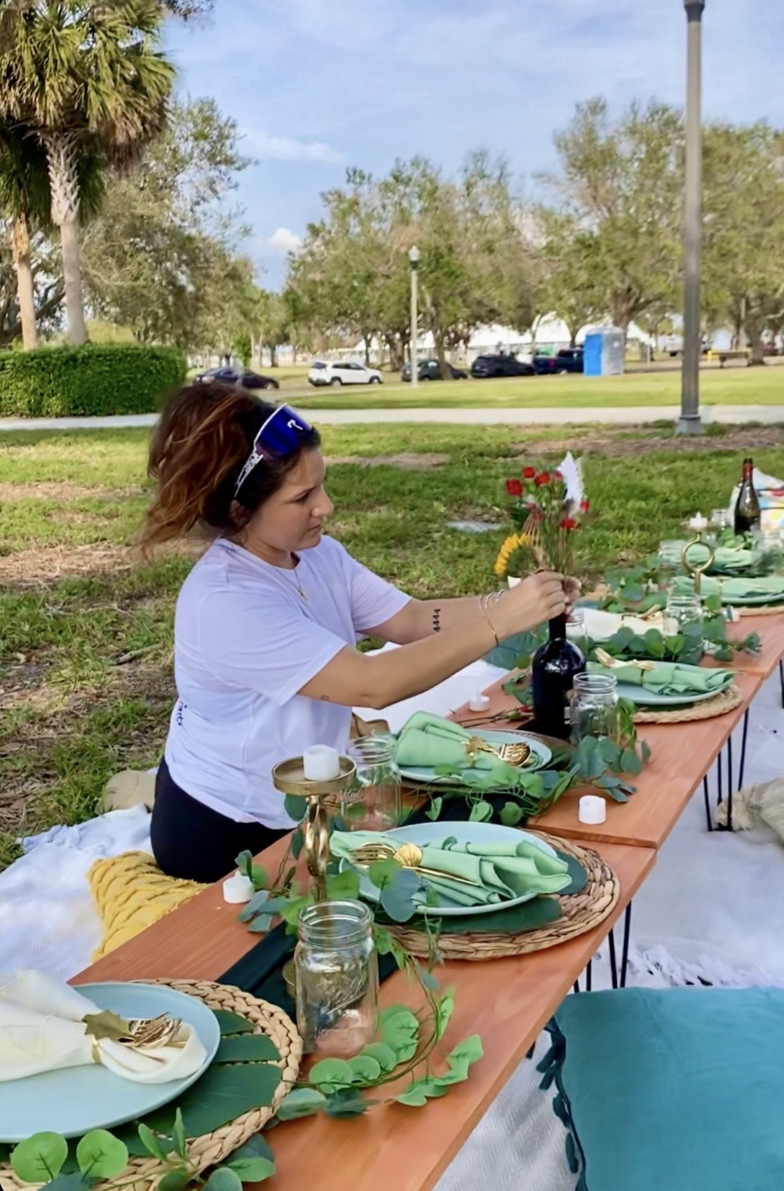 Woman arranging table settings outdoors with plates, glasses, and green napkins, surrounded by trees and parked cars.
