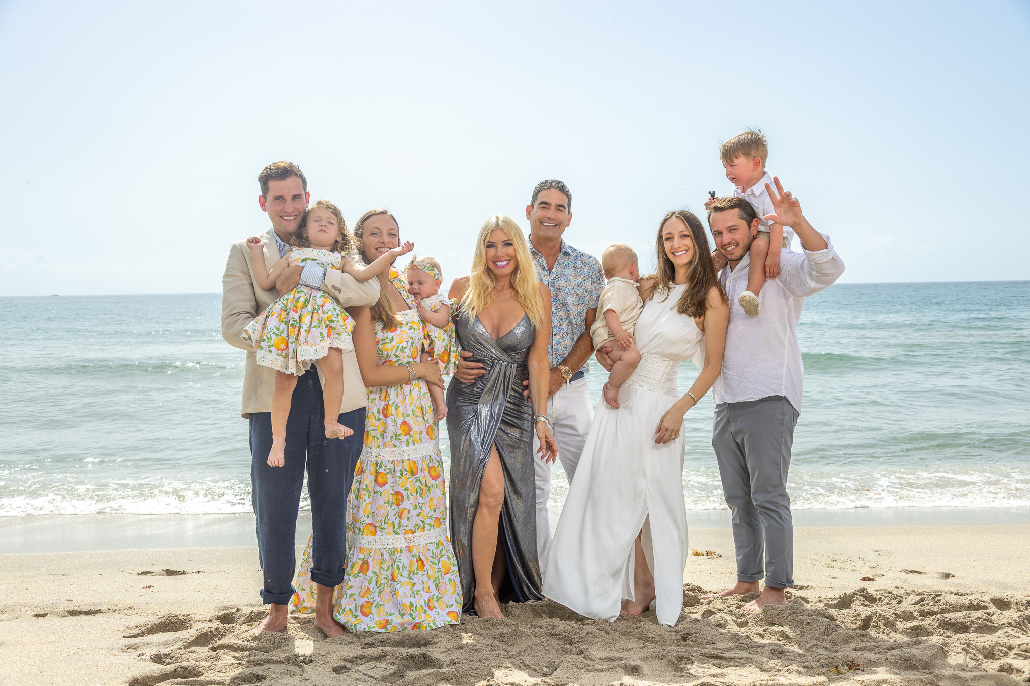 Group of people in wedding attire on a beach, smiling and posing for a photo, ocean in background.