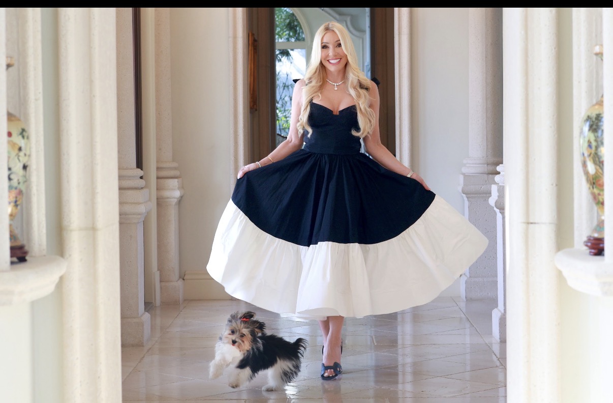 Woman in a navy and white dress standing in a hallway with a small dog, columns on sides, and a window behind.