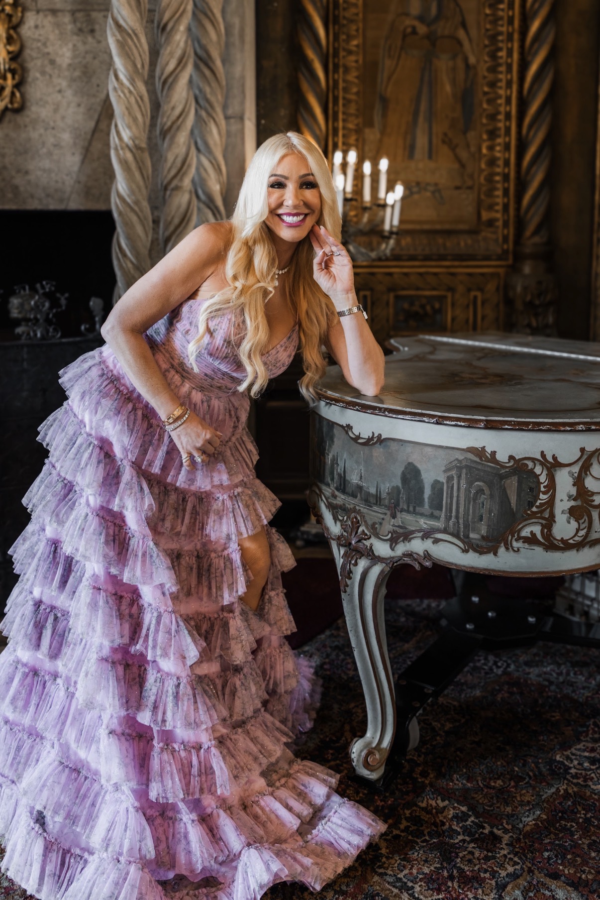 Woman in a purple ruffled gown leaning on a decorated piano in a luxurious room with ornate woodwork and a chandelier.