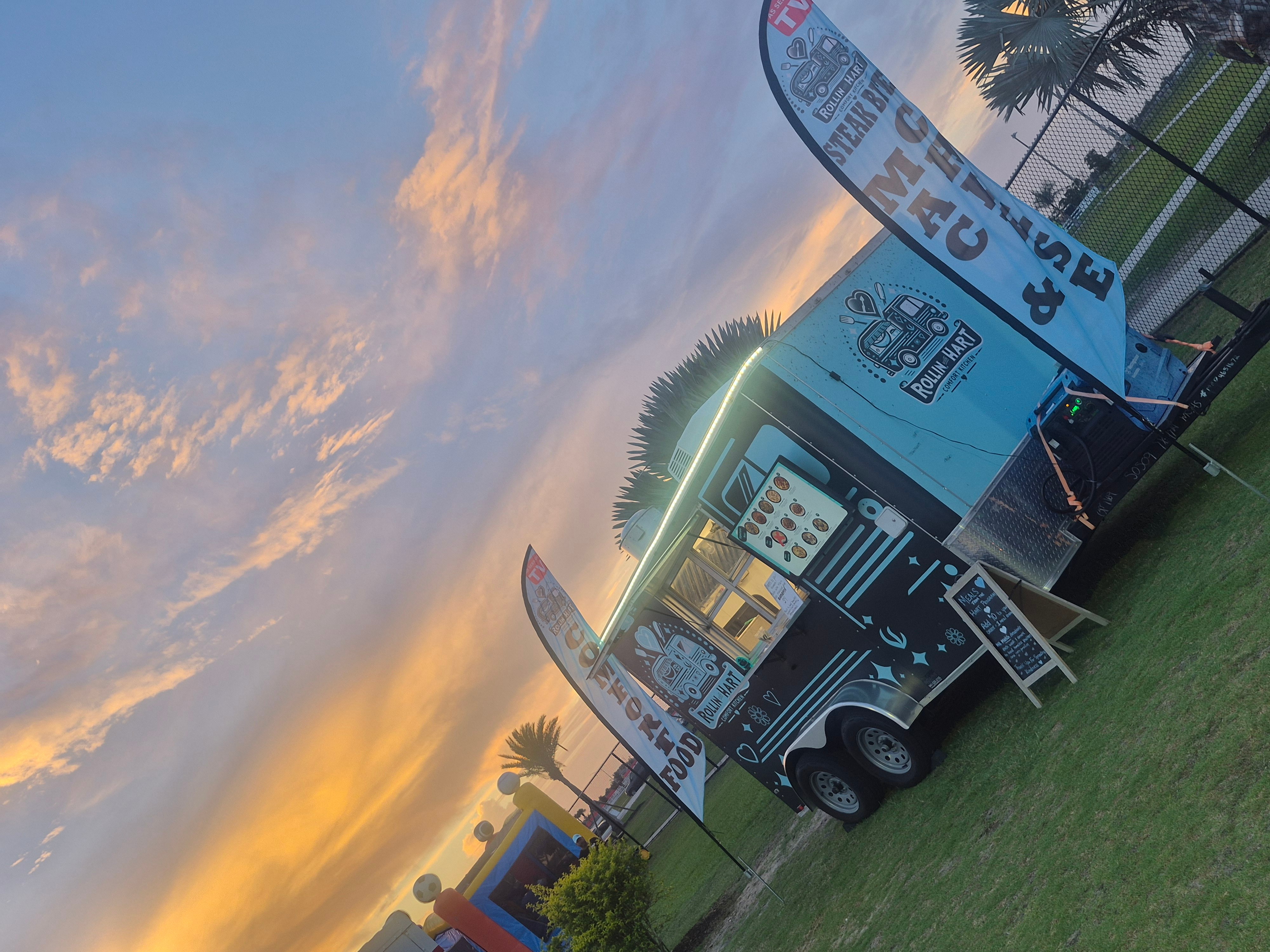 Food truck parked on grass with sunset sky and palm trees in background.