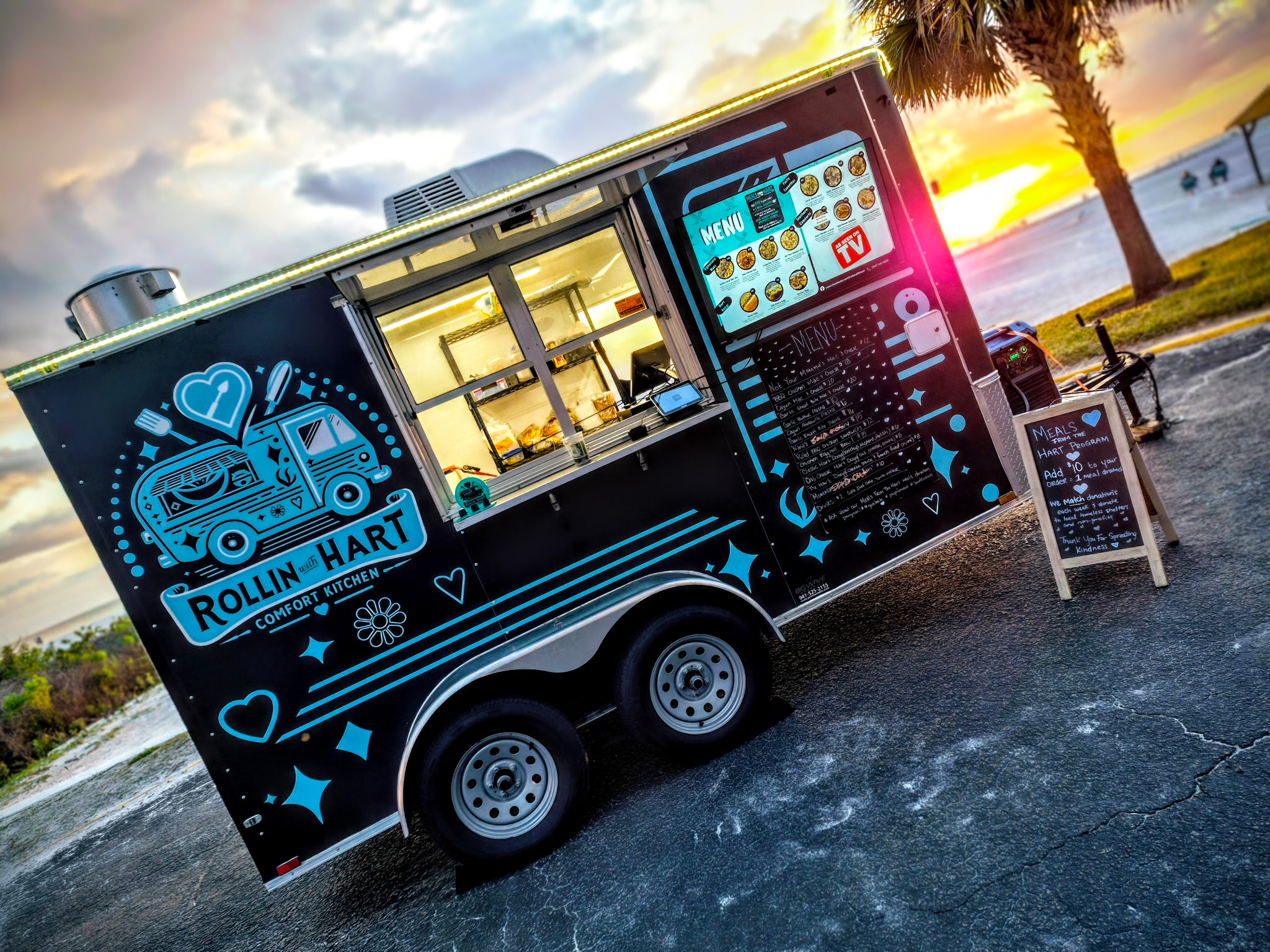 Food truck with digital graphics and a sunset background near a beach, palm trees, and a sidewalk.