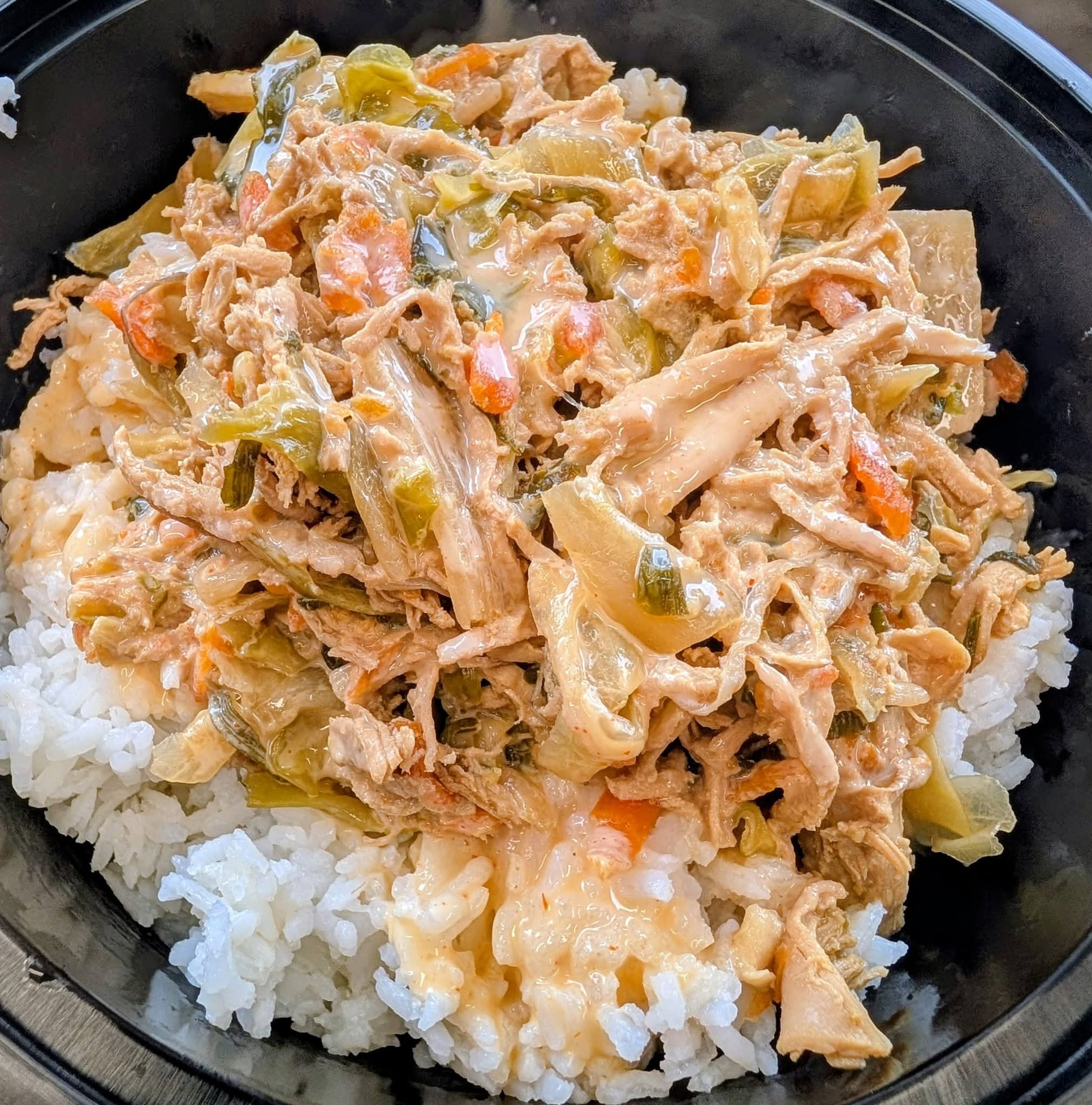 Shredded chicken with vegetables and sauce served over white rice in a black bowl.