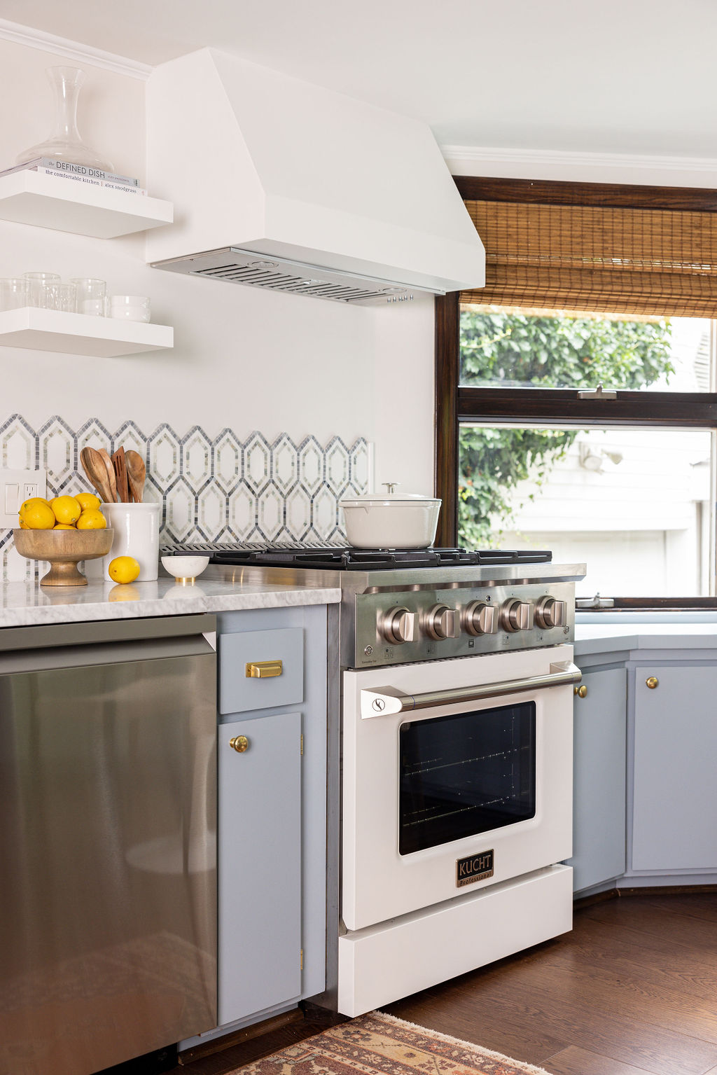 Kitchen with stove, blue cabinets, white range hood, window with bamboo shade, and countertop with lemons and utensils.