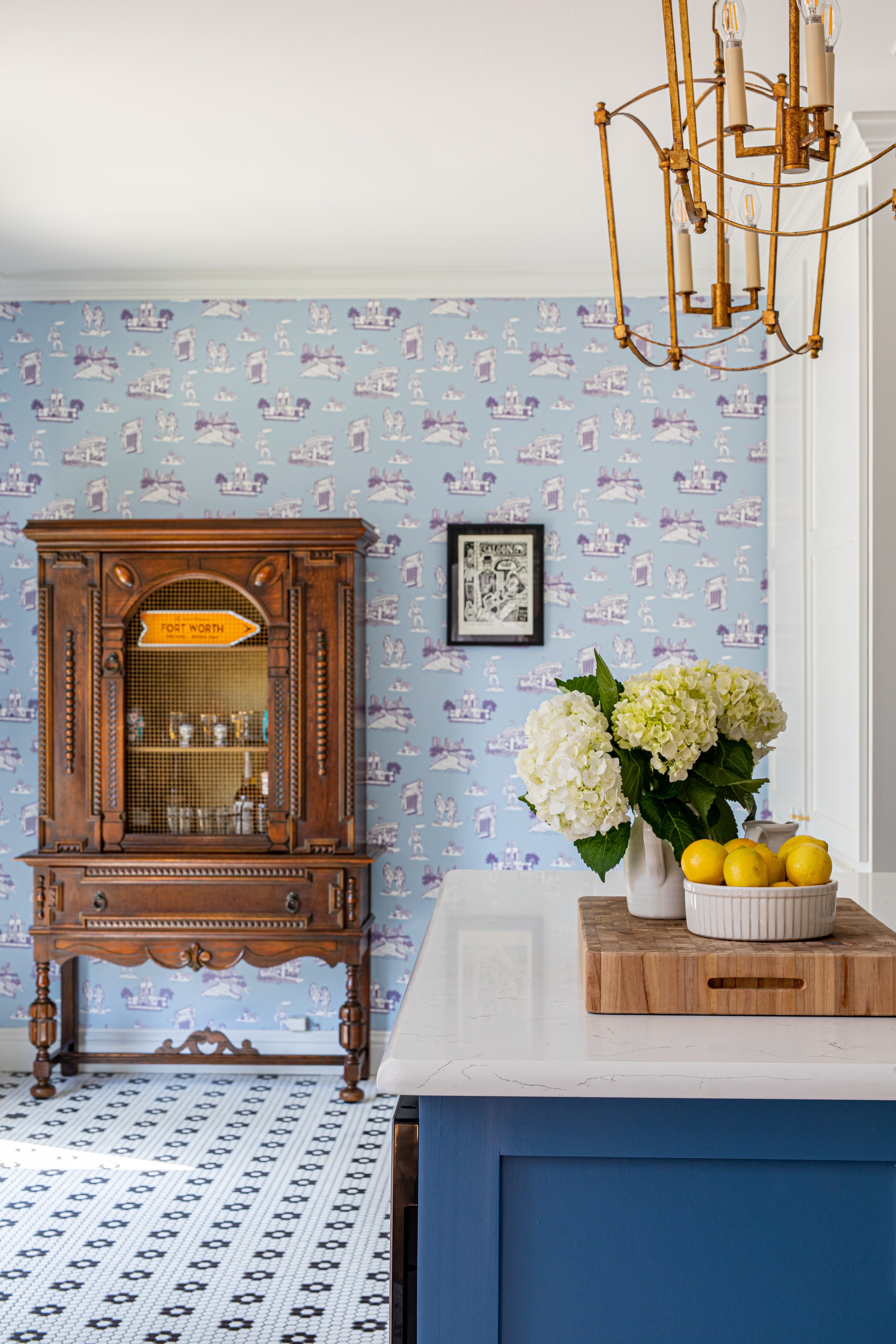 Interior room with patterned wallpaper, wooden cabinet, white flowers, and a bowl of lemons on a blue counter.