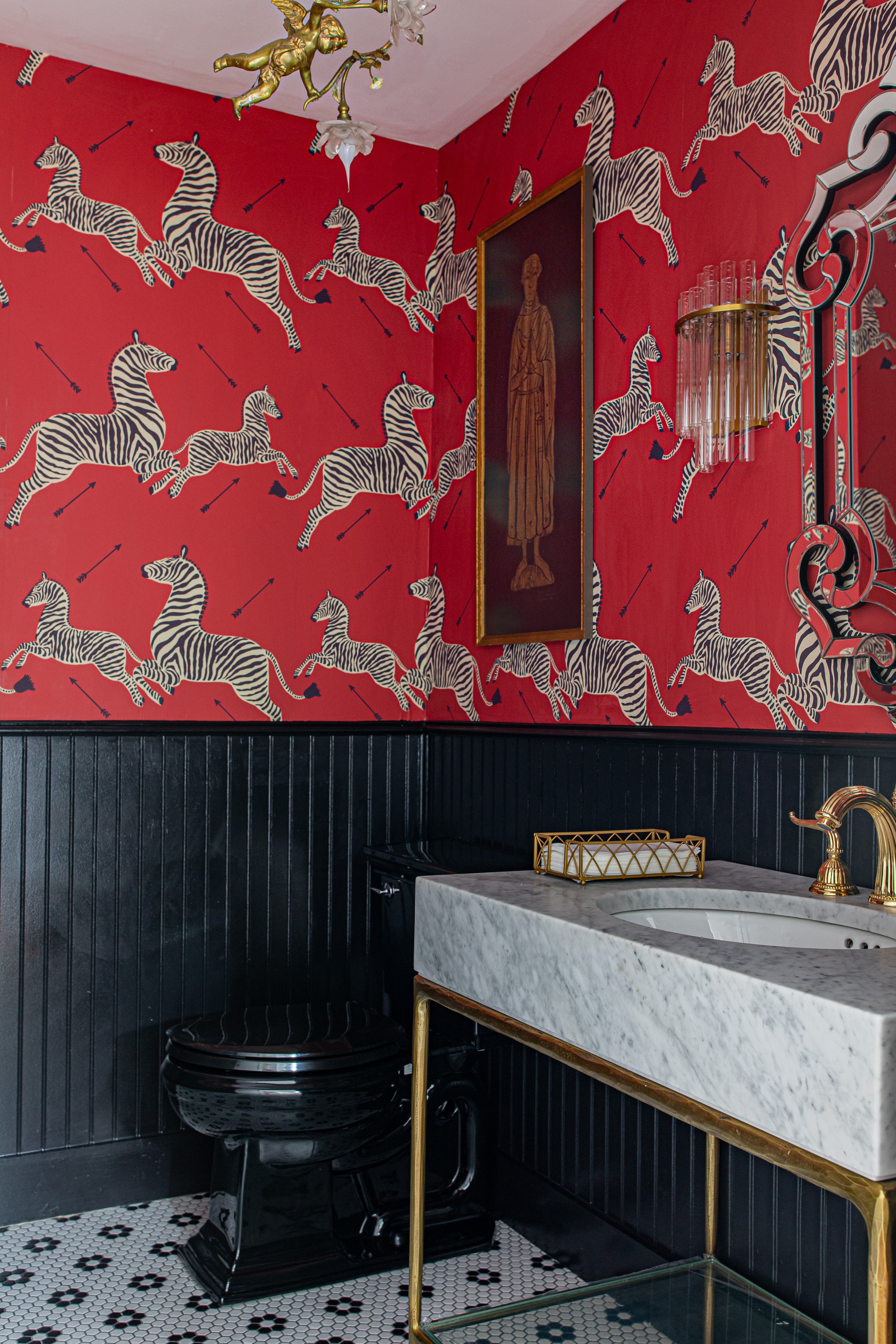 Red wall with zebra wallpaper, framed artwork, black wainscoting, marble sink, gold fixtures, and a black stool.