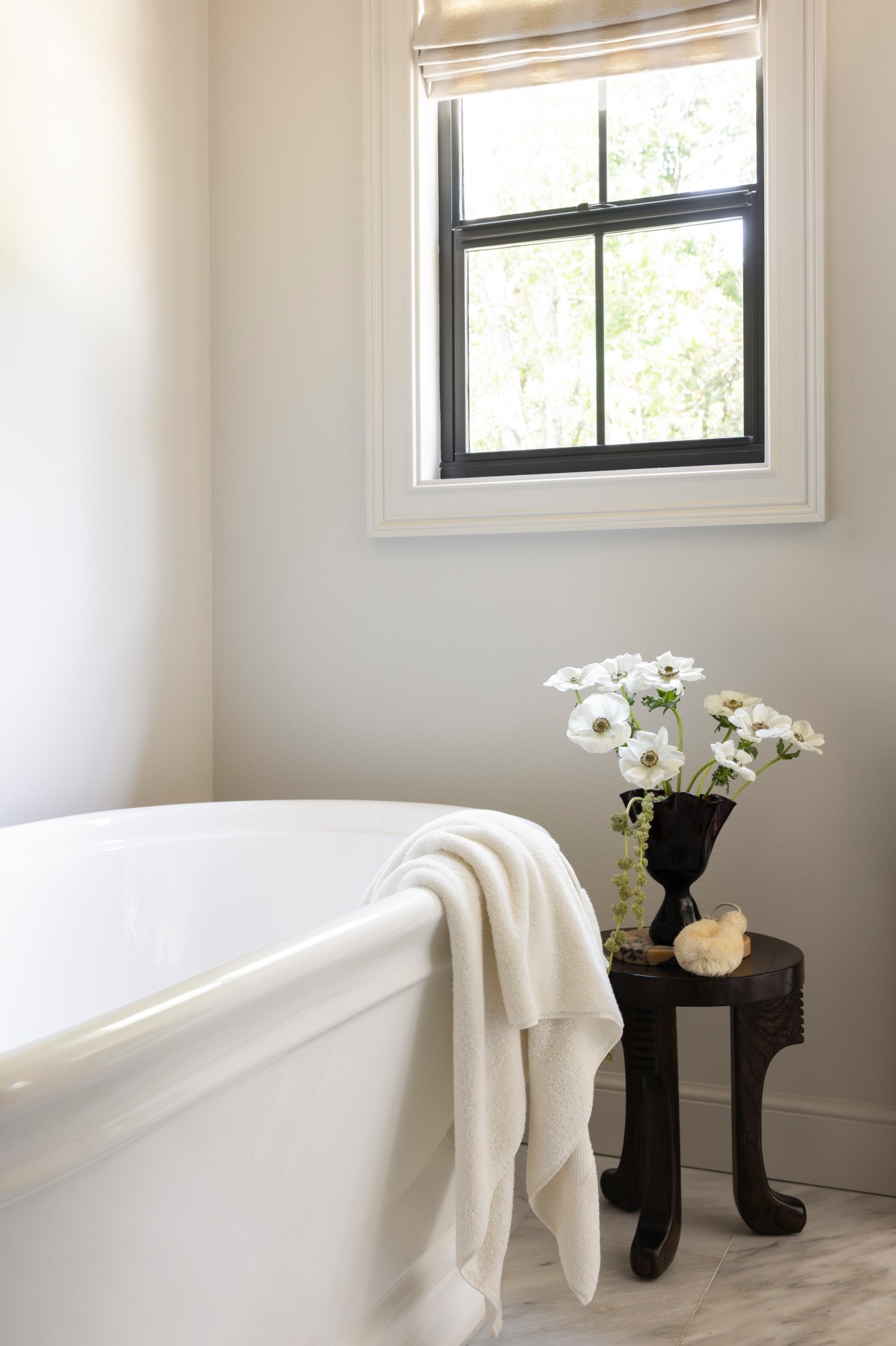 Bathroom corner with a white bathtub, a black side table with white flowers, and a window with a view outside.