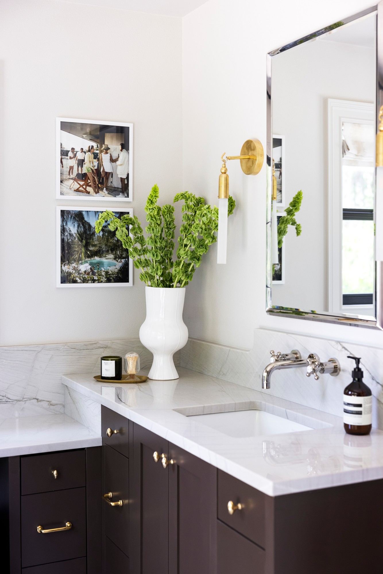 Bathroom vanity with a white vase holding green plants, a mirror, and framed photos on the wall.