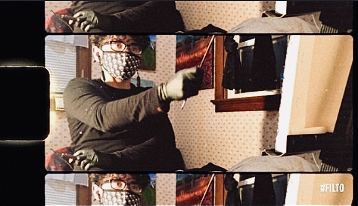 Person wearing glasses, a face mask, and gloves, pointing at a wall with artwork, in a room with patterned wallpaper.