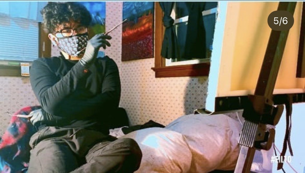 Person wearing glasses and a face mask, sitting indoors with a computer and a person lying down nearby.