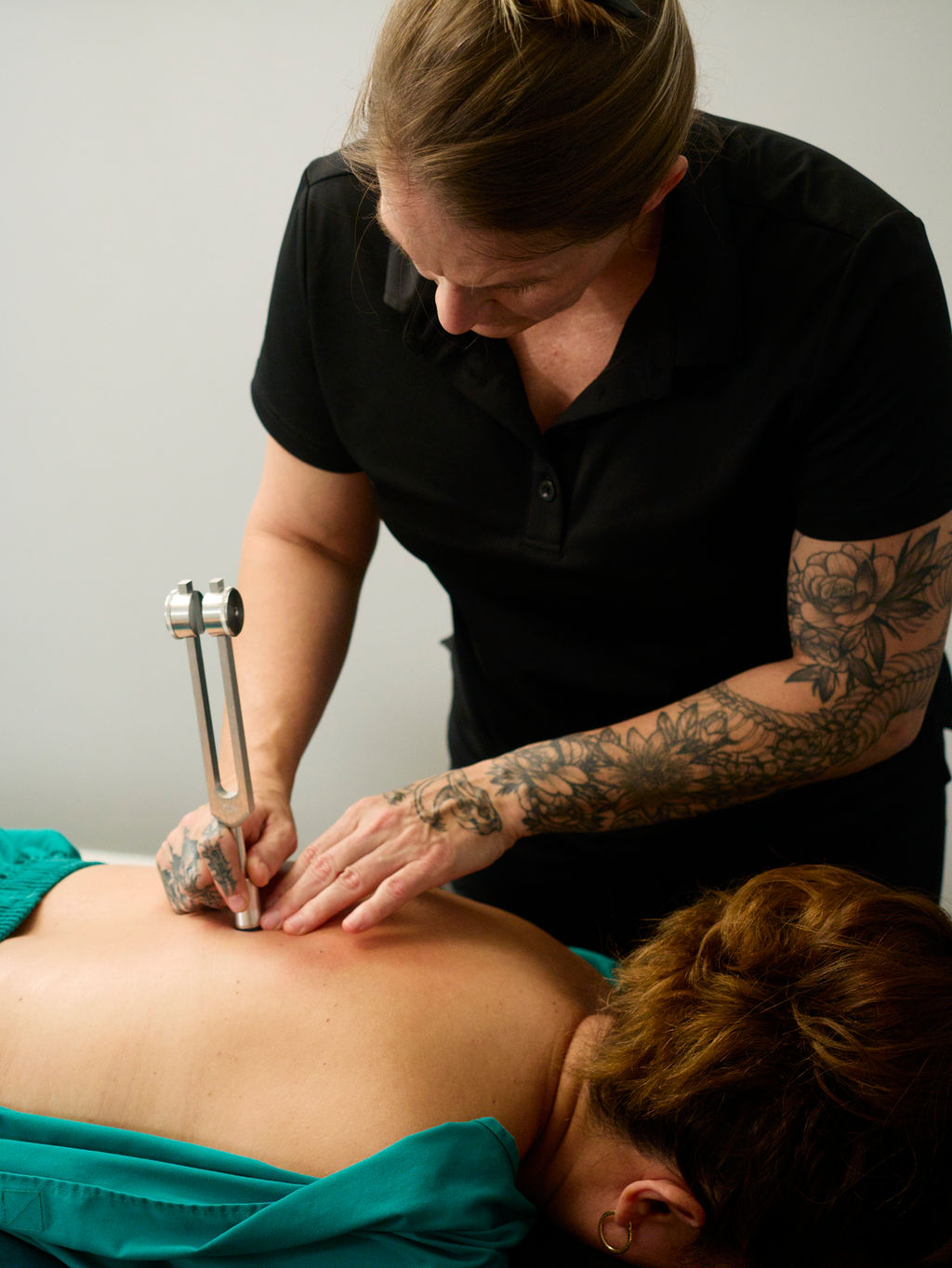 Tattooed woman performs a procedure on a person's back using a handheld device, in a clinical setting.