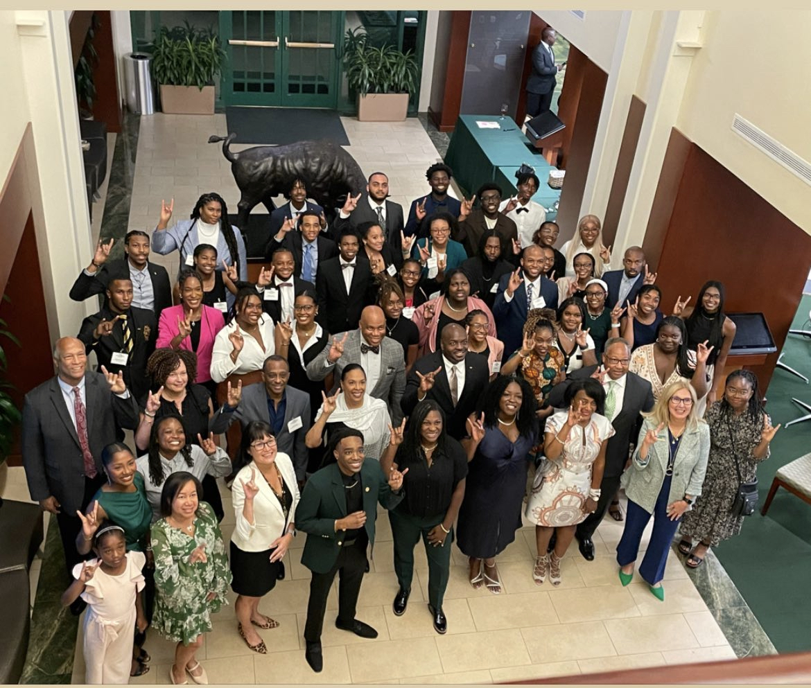 Group of diverse people gathered indoors, smiling and waving at the camera, in a spacious lobby area.