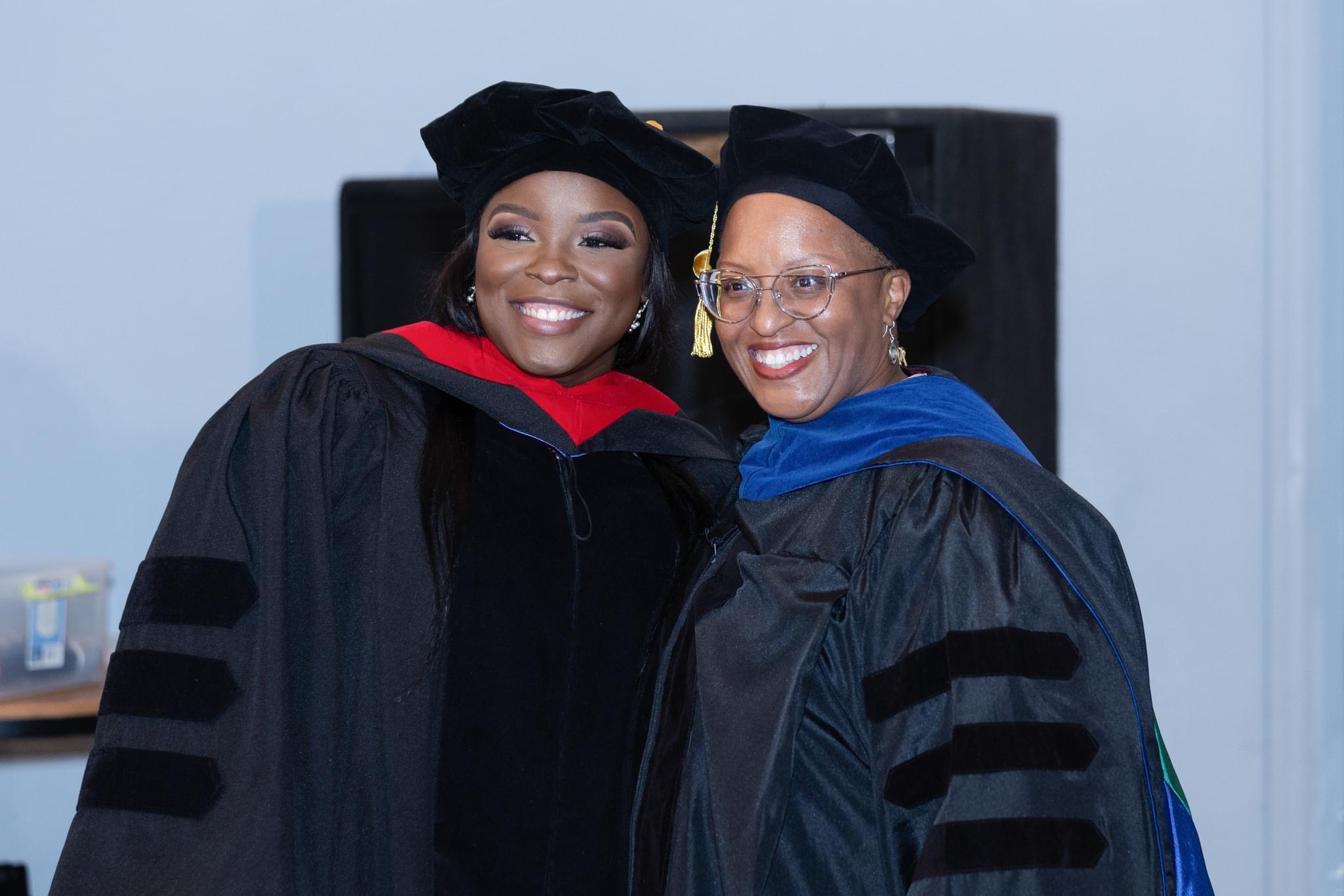Two women in graduation gowns and caps smiling indoors, one with glasses and a blue stole, the other with a red collar.