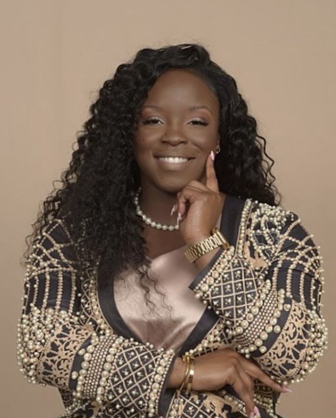 Woman with curly hair smiling, wearing a patterned jacket, pearl necklace, and gold watch, posing with her hand near her face.