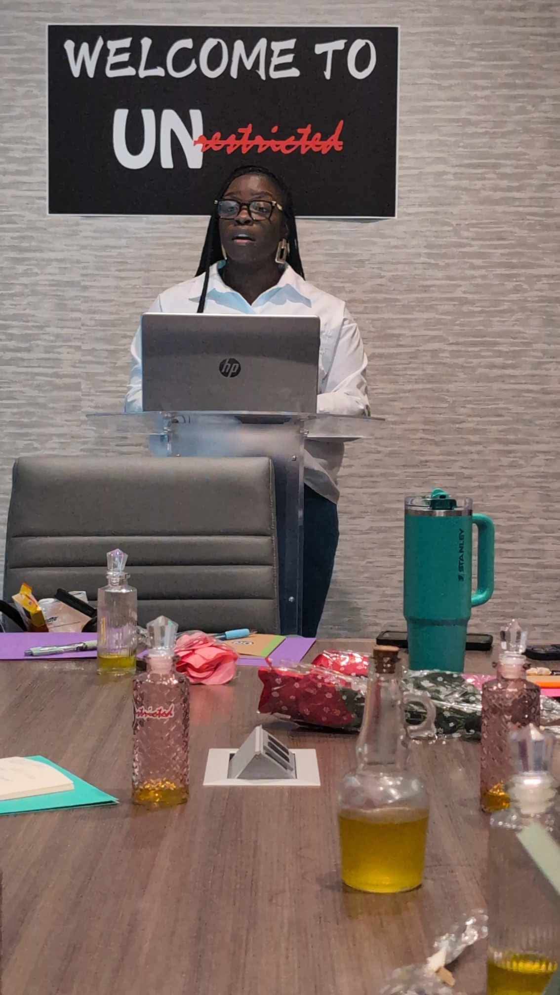 Person standing behind a table with a laptop, in front of a sign that says 'WELCOME TO UN', with various bottles and items on the table.