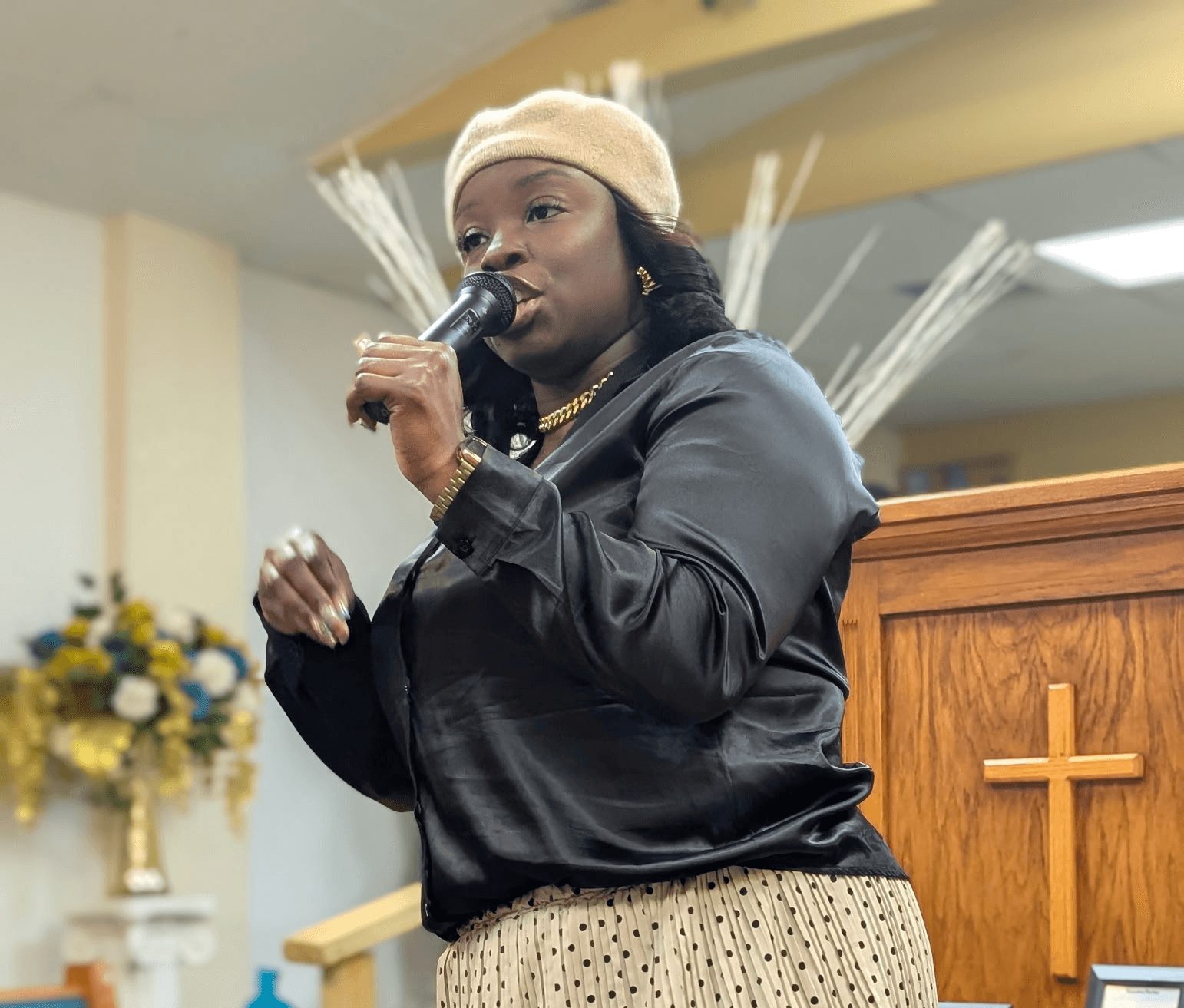 Woman speaking into microphone in a church setting, wearing a beige hat and black jacket, with a wooden cross behind her.