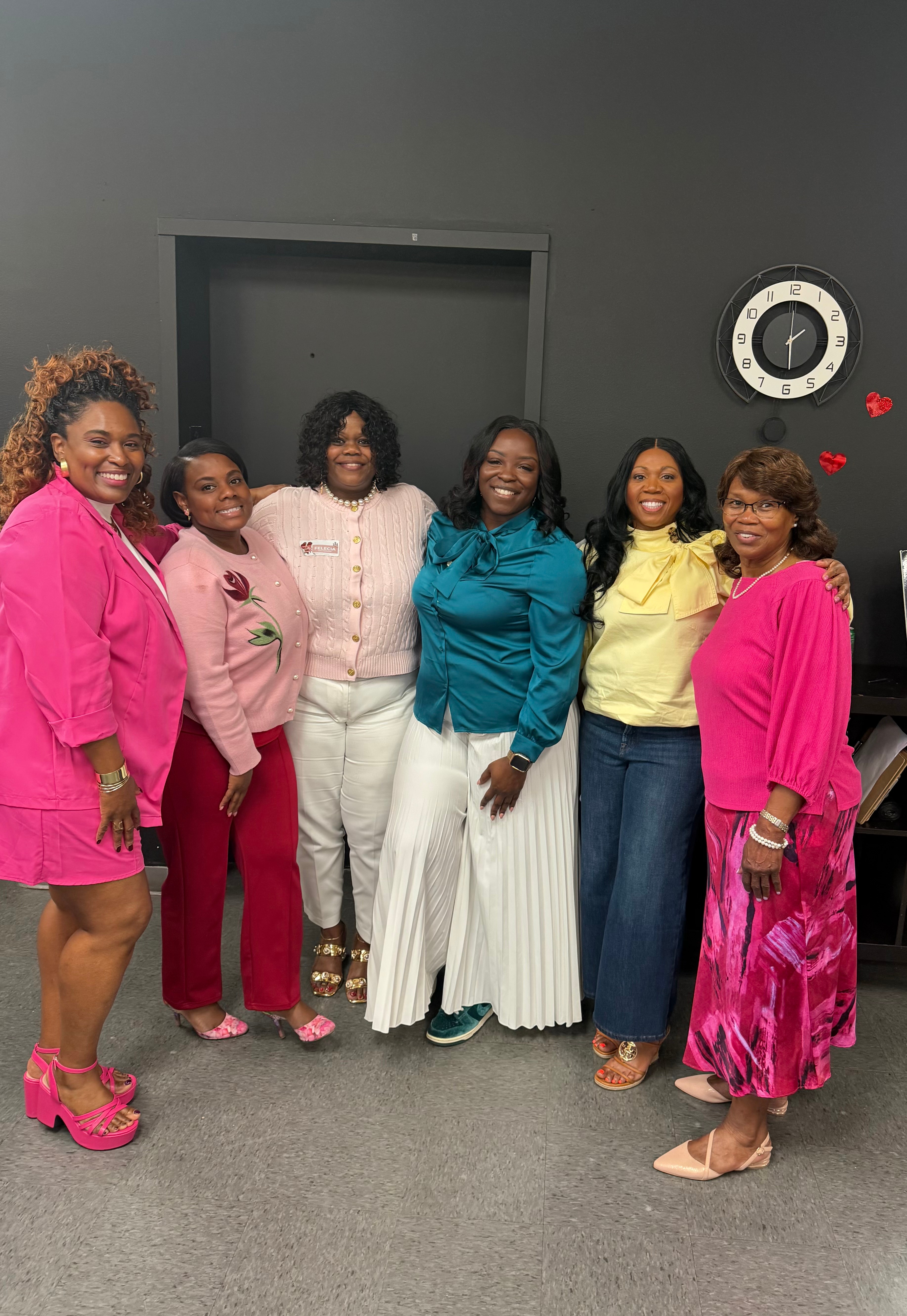 Six women standing together in a room, smiling, wearing colorful outfits, with a dark wall and clock behind them.